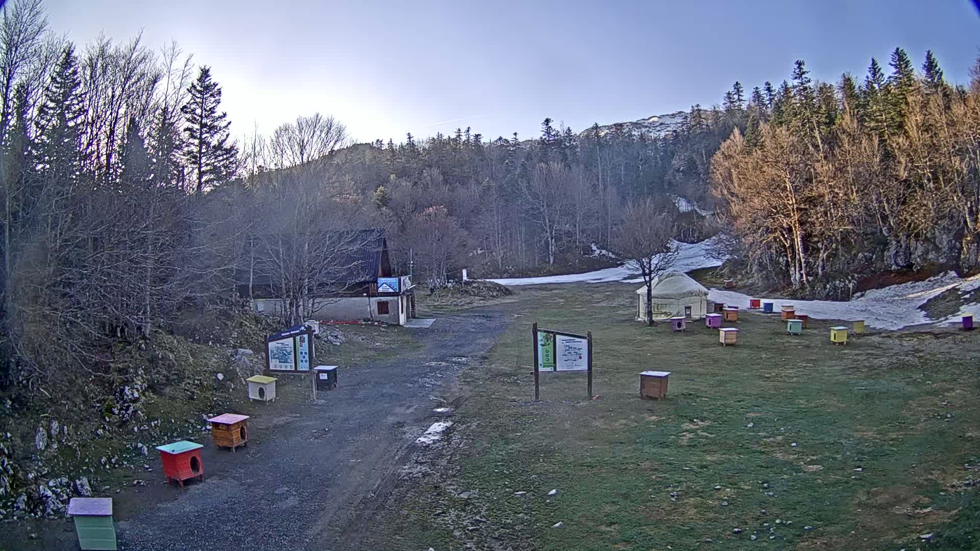 A small building sits near a mostly cleared area with patchy snow, surrounded by leafless trees and colorful small structures on a clear, bright day.