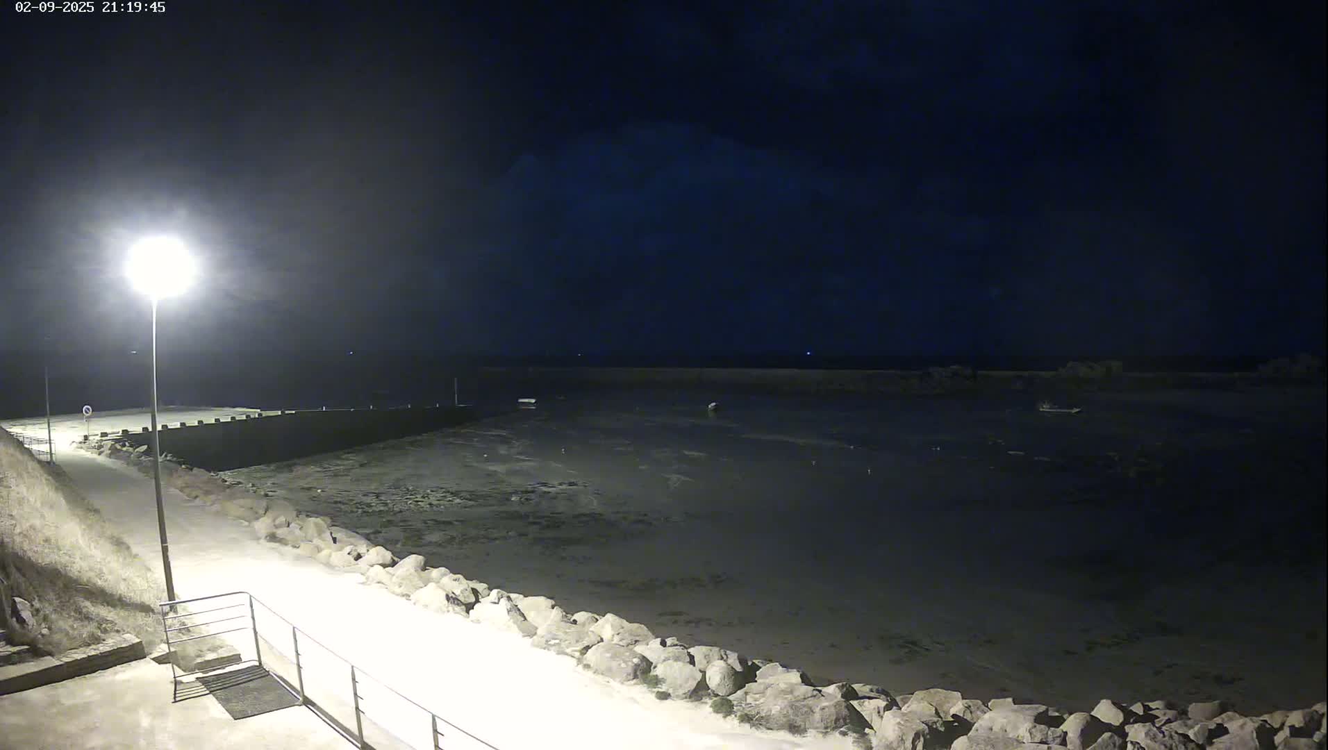 A nighttime view of a harbor with a few boats, a rocky shoreline, and a lit walkway.