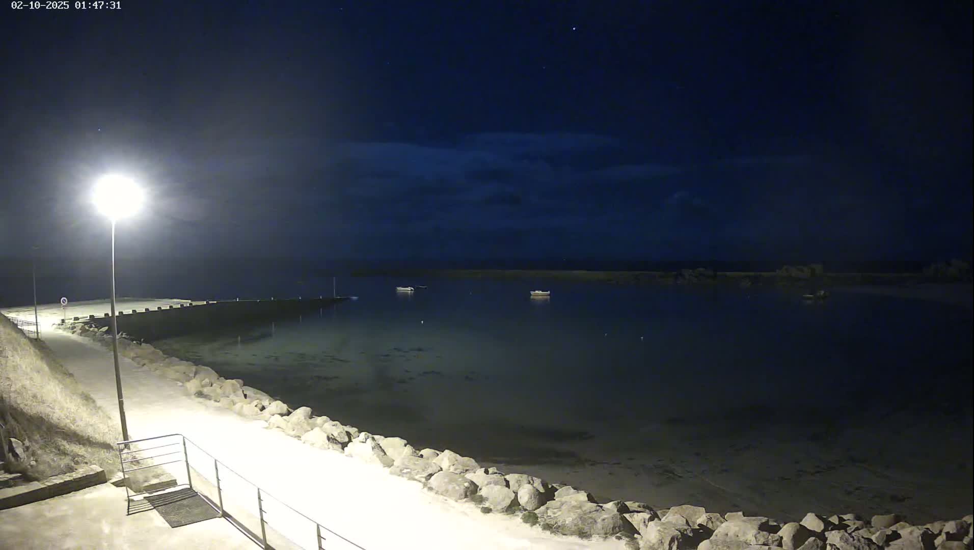 A nighttime view of a calm body of water with a few boats, a lit pathway along a rocky shoreline under a dark, partly cloudy sky.