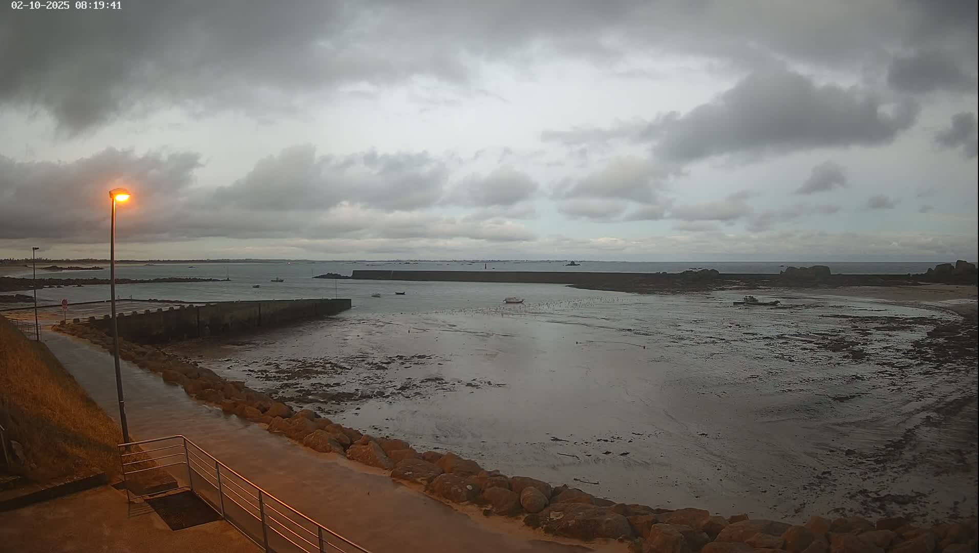 A low-tide harbor scene under a cloudy sky shows a paved walkway leading to a harbor wall, with several boats visible in the shallow, muddy water.