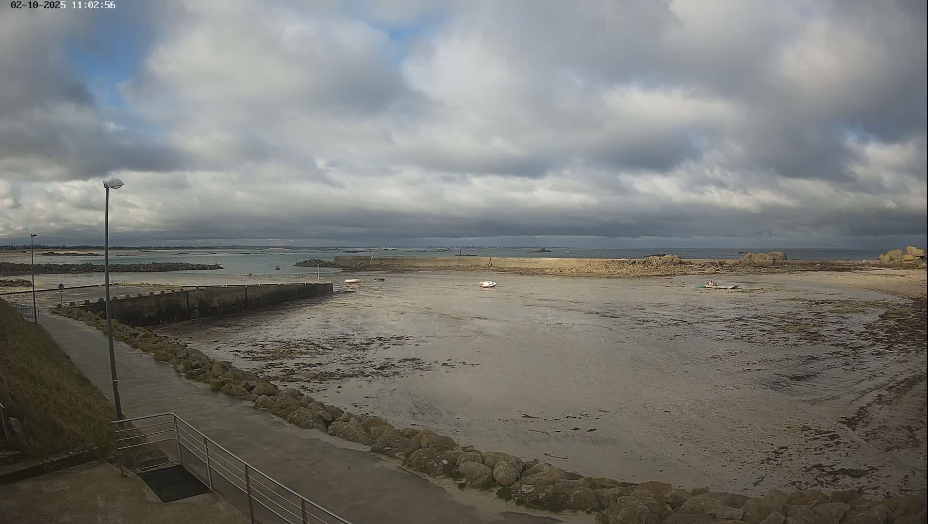 A mostly cloudy day reveals a low-tide harbor scene with several small boats resting on exposed mudflats, surrounded by rock walls and a paved walkway.