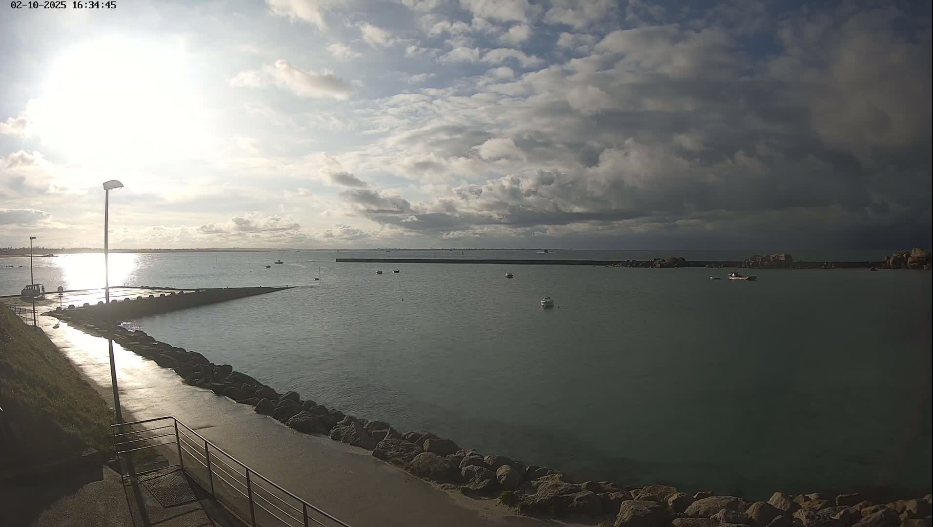 A partly cloudy sky illuminates a calm harbor with several small boats, a stone walkway, and a breakwater under a mostly cloudy sky.