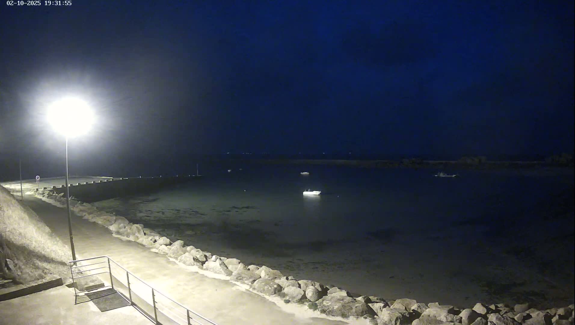 A nighttime view of a calm harbor with a few boats, a rocky shoreline, and a lit walkway.
