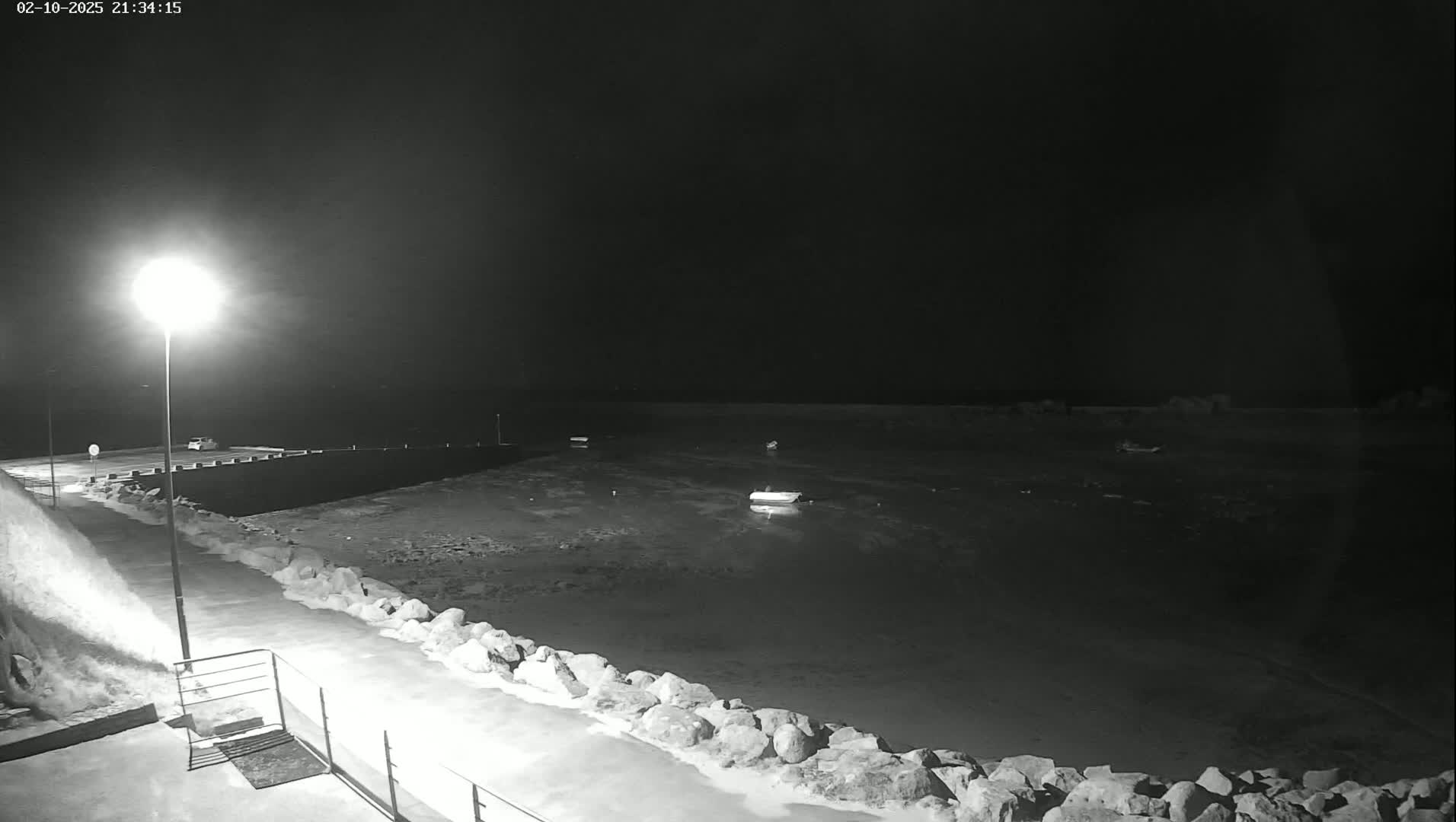 A nighttime, low-light view of a small harbor with several boats, a seawall lined with rocks, and a single light illuminating a paved area.