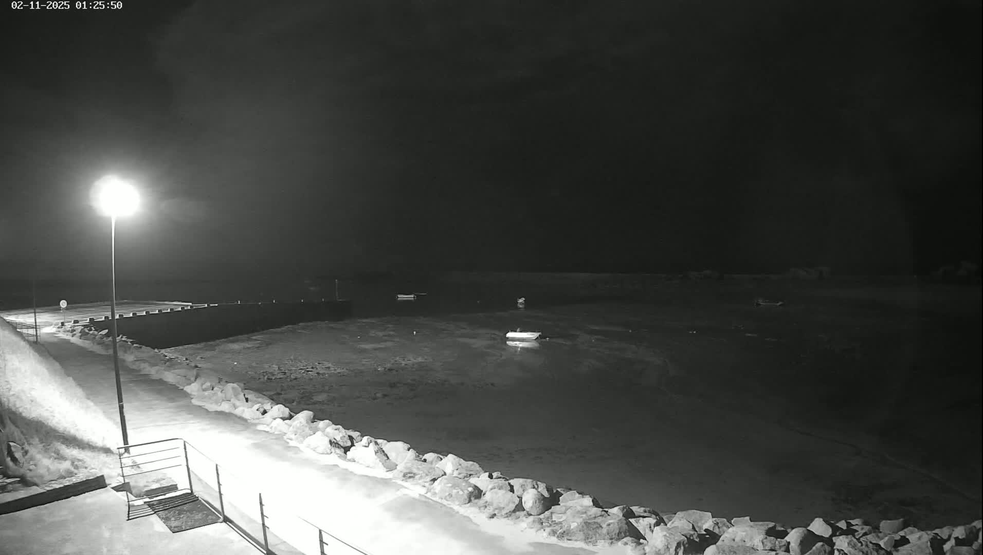 A nighttime, black-and-white view of a calm harbor with several small boats moored, illuminated by a single streetlight, and a rocky shoreline partially covered in snow or ice.