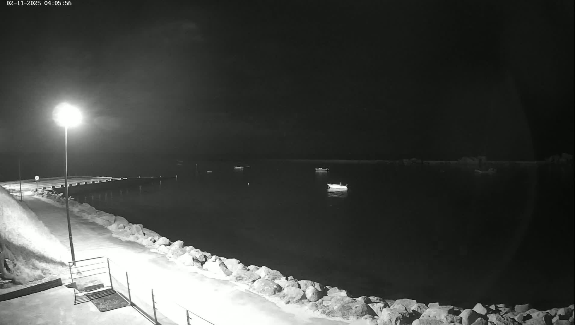 A nighttime view of a calm body of water with several small boats, a lit pier, and a snow-covered rocky shoreline under a dark, cloudy sky.