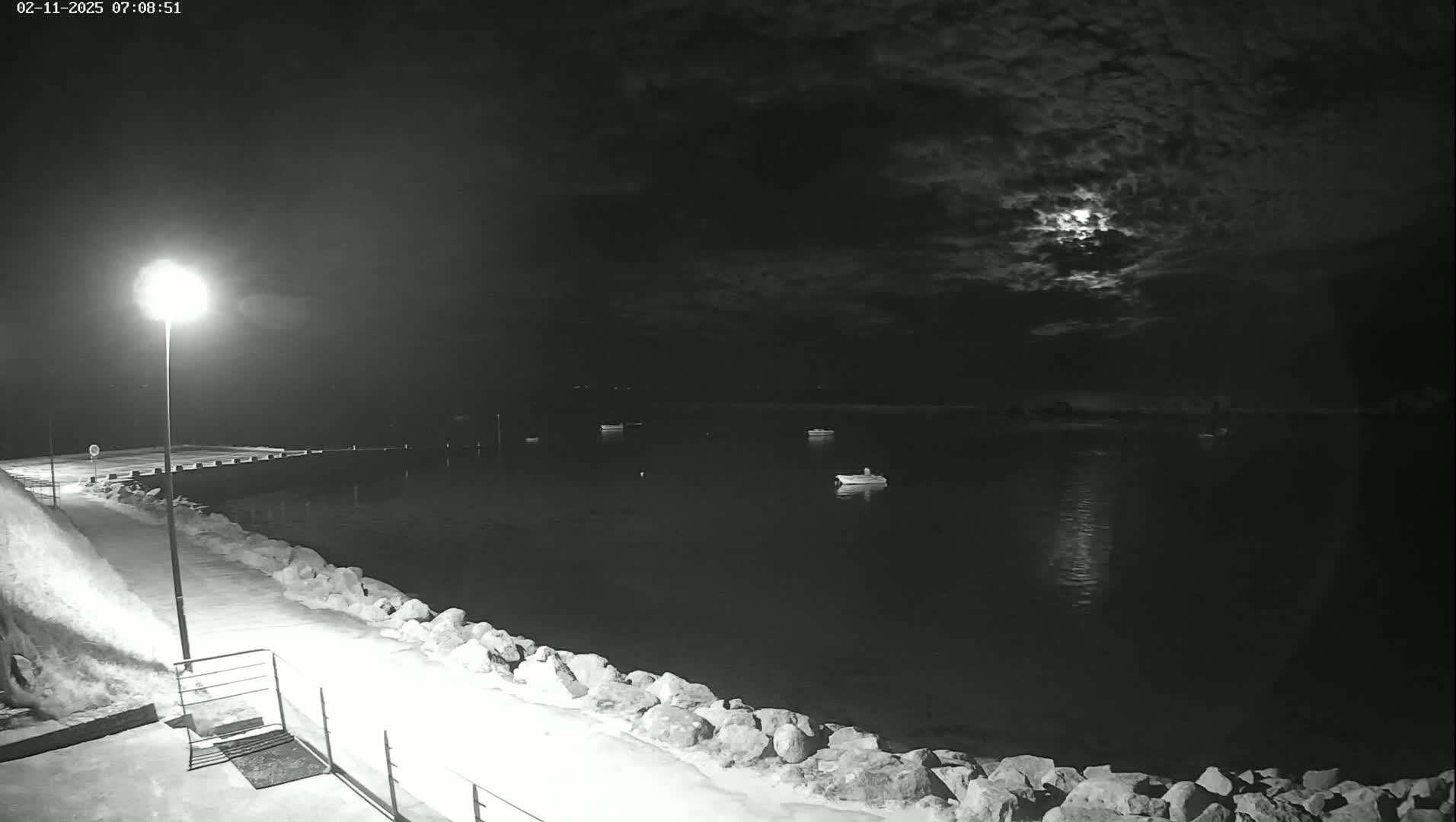 A moonlit night scene shows a snow-covered shoreline with a few boats in the calm water, lit by a lamppost on the walkway.