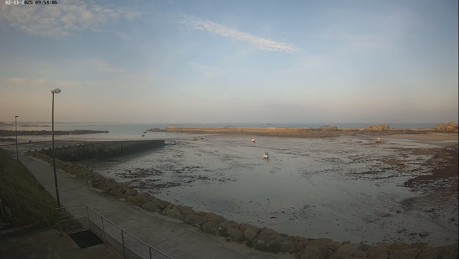 A mostly clear, bright day shows a harbor with several small boats resting at low tide, a stone jetty, and a paved walkway.