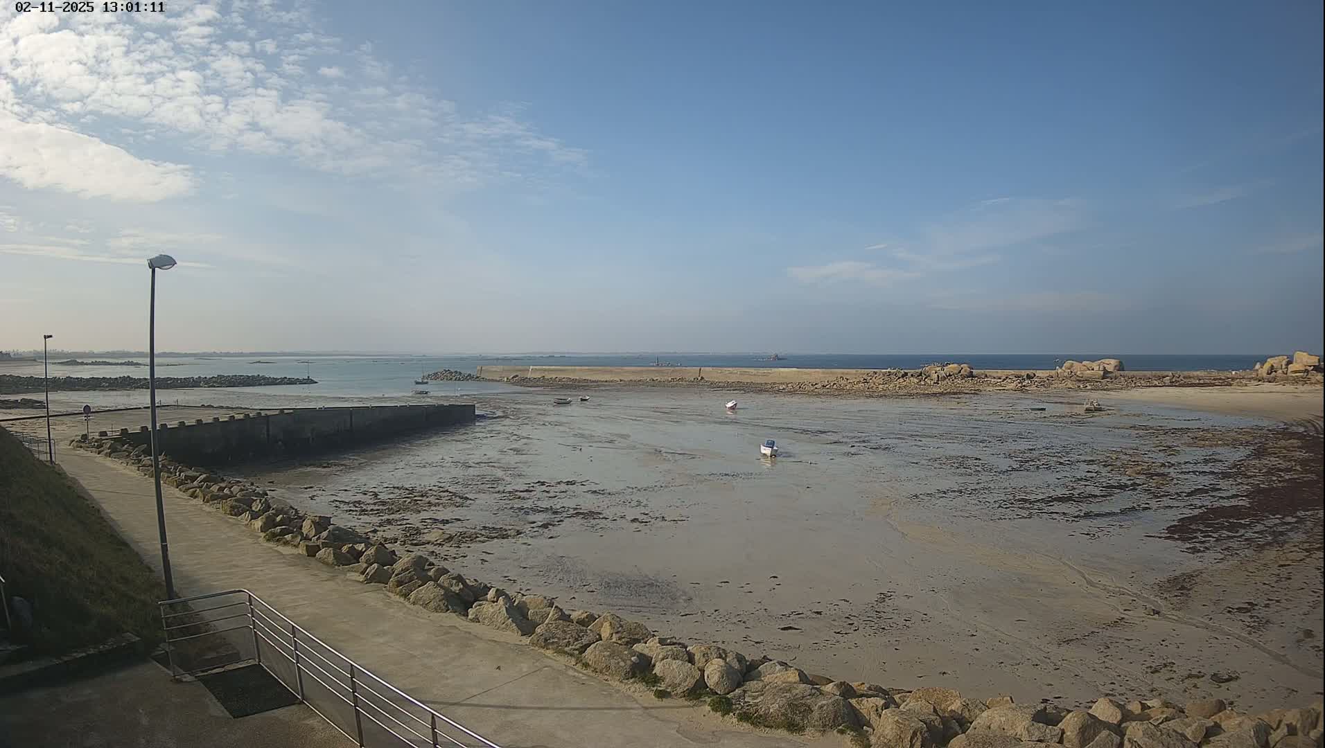 A mostly sunny day reveals a harbor with a low tide exposing a wide expanse of mudflats and several small boats, a walkway leads toward the water's edge.