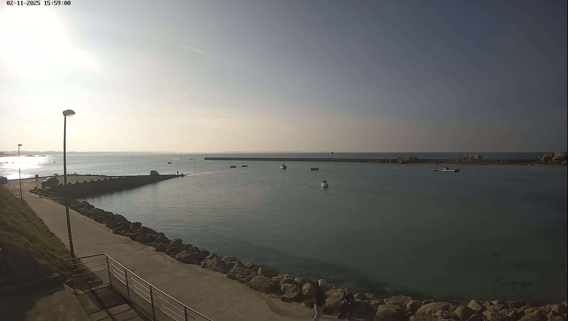 A calm, sunny day at a harbor shows several small boats in the water, a paved walkway leading to a stone jetty, and a few people walking near the water's edge.