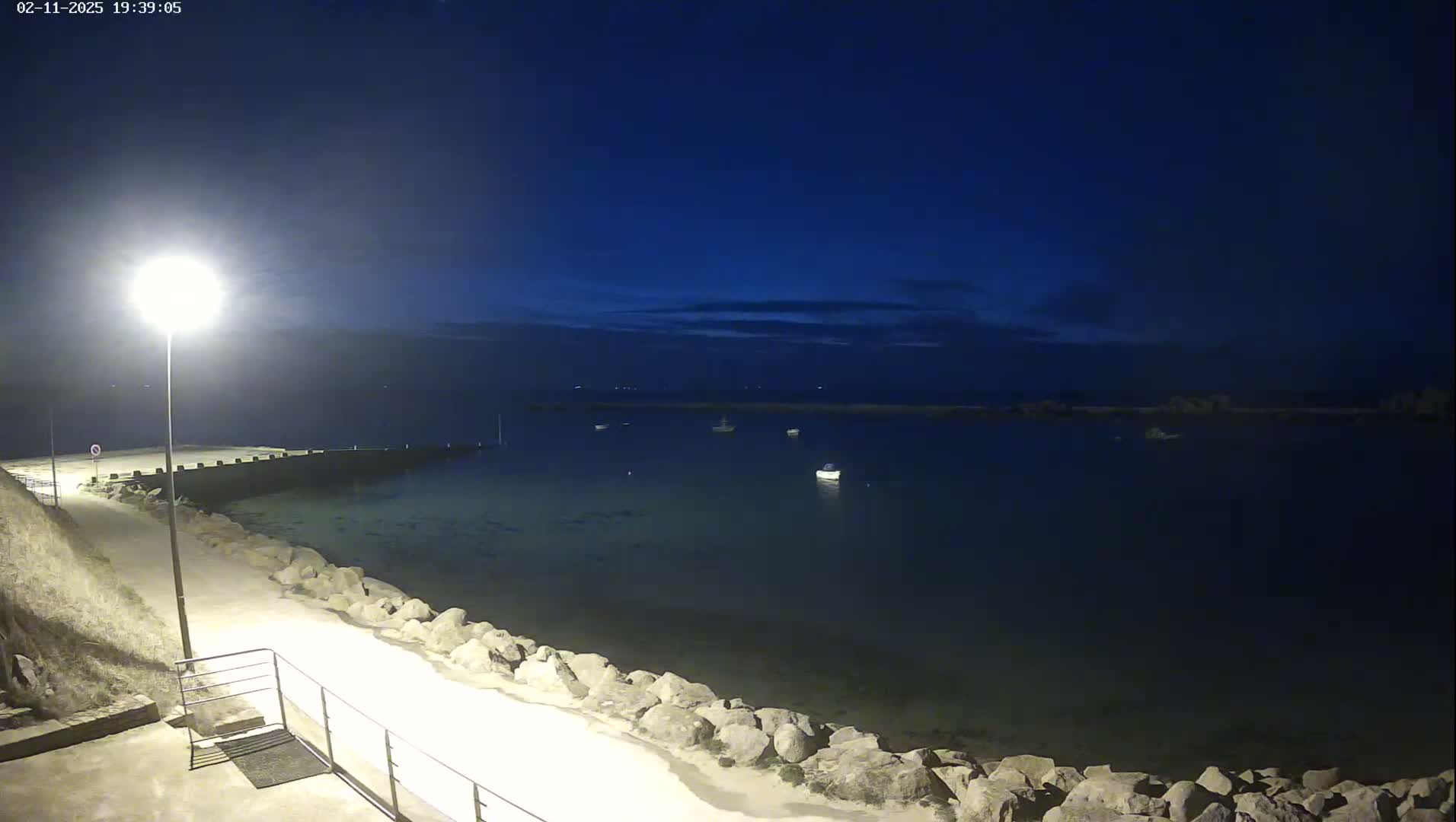 A nighttime view of a calm body of water with several boats, a stone seawall, and a paved walkway lit by a single lamppost.