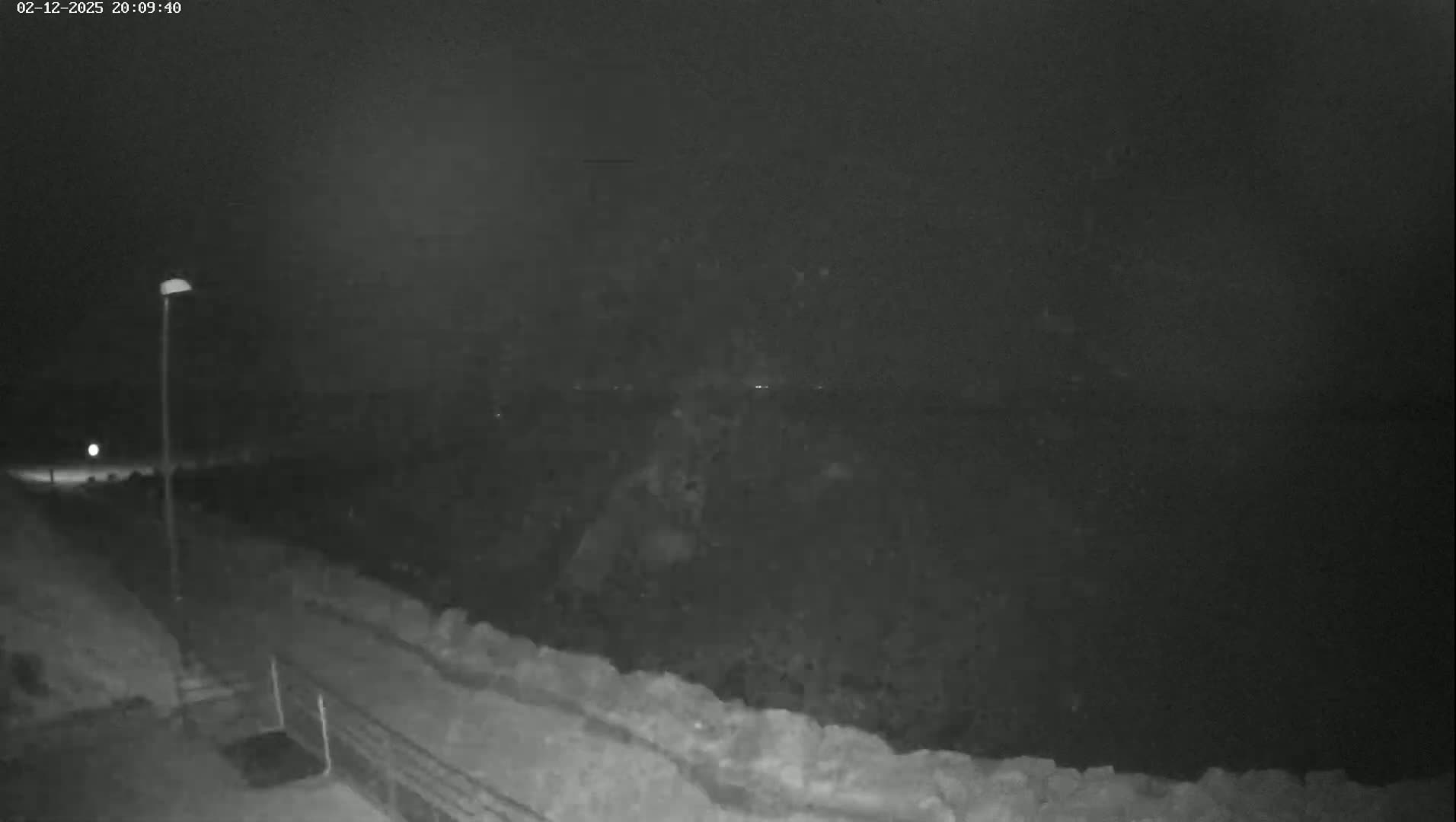 A nighttime, low-light view shows a paved path next to a low stone wall, extending toward a dark body of water under a mostly clear sky.
