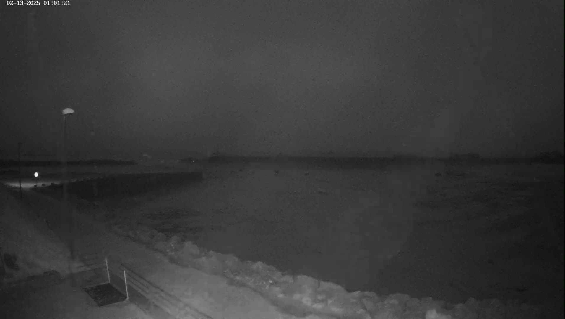 A nighttime, snowy scene shows a dark body of water behind a snow-covered embankment with a single lamppost.