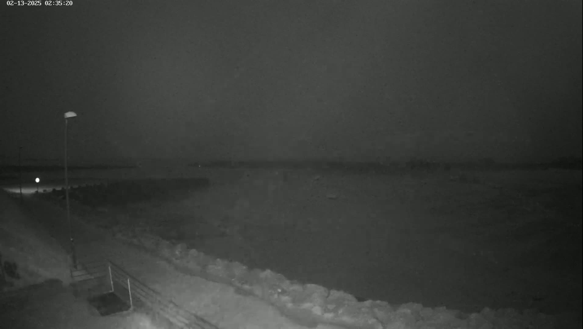 A nighttime, snowy scene shows a dark body of water behind a snow-covered walkway and seawall under a dark, overcast sky.