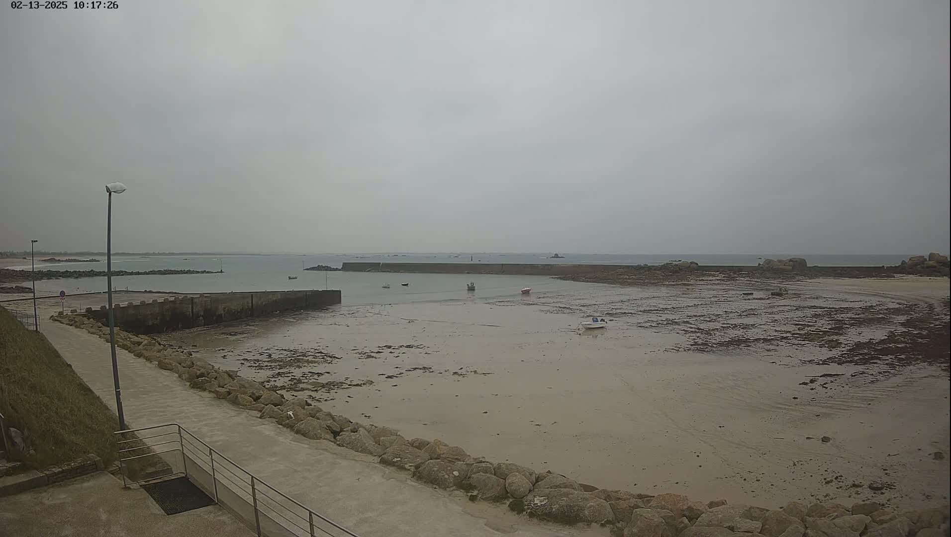 A mostly overcast day reveals a small harbor with several small boats at low tide, surrounded by rocks and a paved walkway.