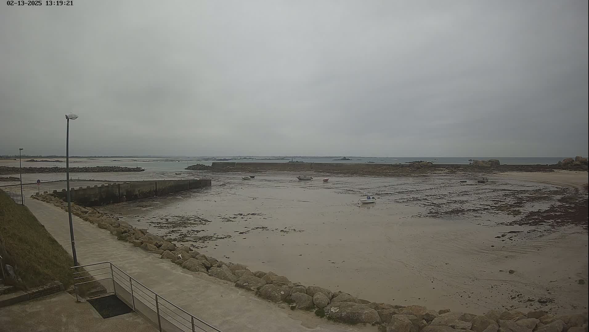 A low-tide, overcast day reveals a small harbor with several small boats resting on the exposed seabed, a stone wall enclosing the harbor, and a paved walkway leading to it.