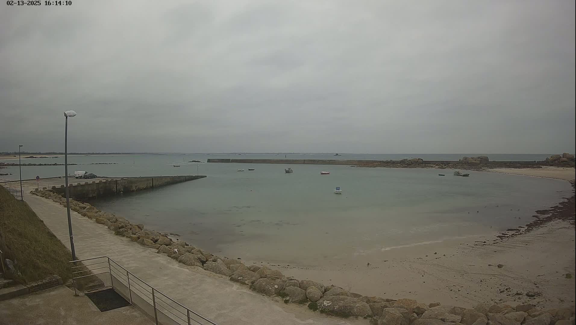 A mostly calm, light-grey body of water is enclosed by a stone jetty, with a sandy beach and several small boats visible under an overcast sky.