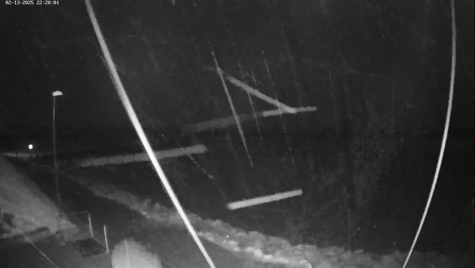 A nighttime, snowy scene shows a road, a lamppost, and some indistinct objects in the middle ground.
