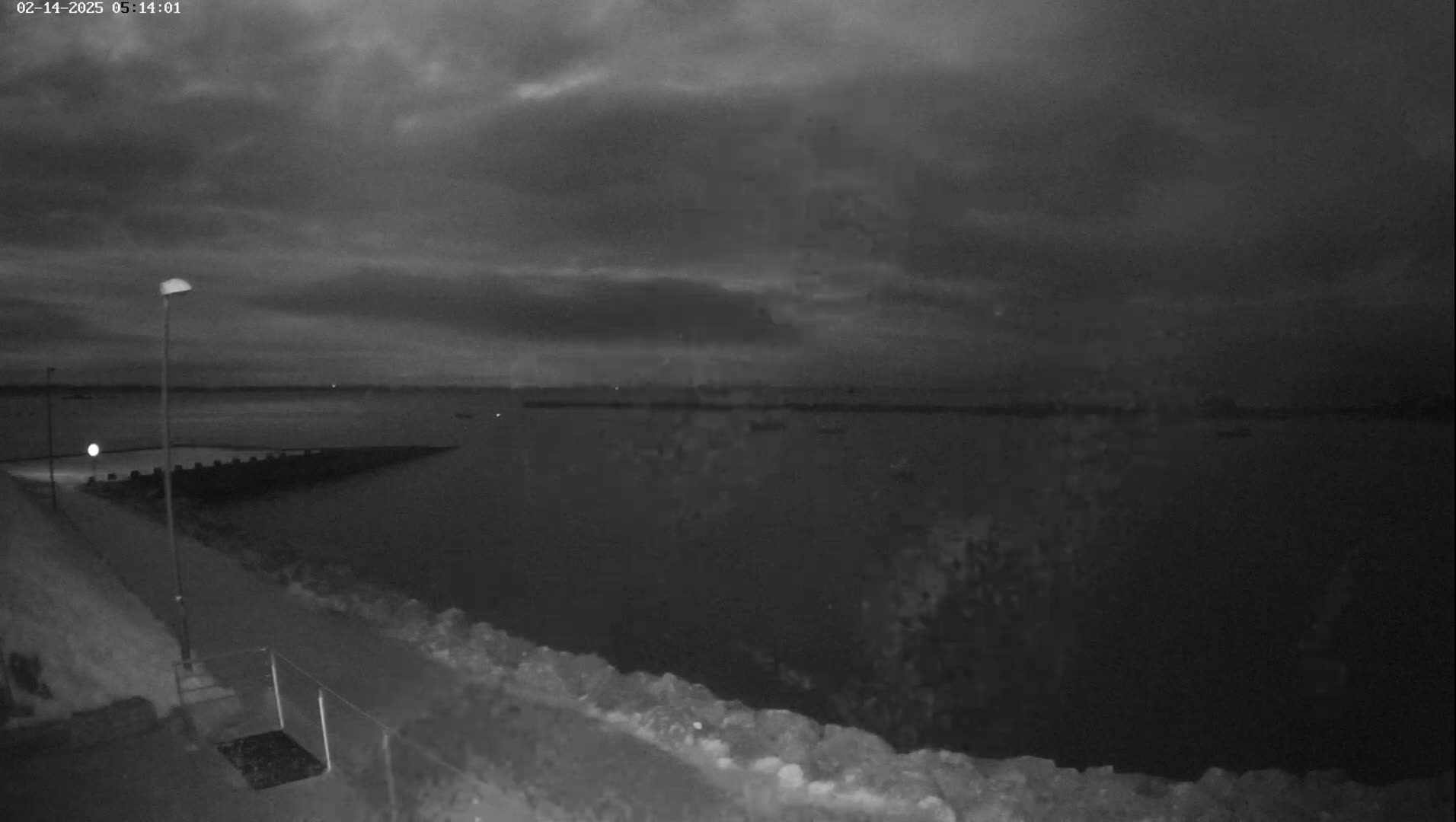 A nighttime view of a calm body of water under a dark, cloudy sky, shows a shoreline with a lamppost and a small structure.