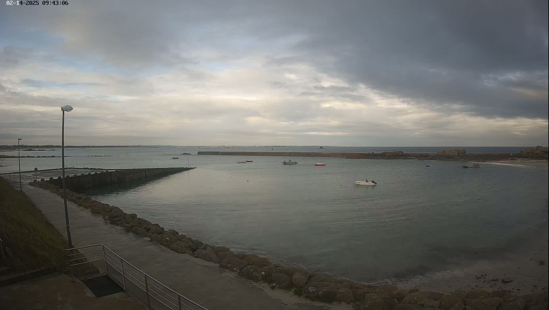 A calm, overcast day shows a small harbor with several small boats moored, a paved walkway, and a rocky shoreline.