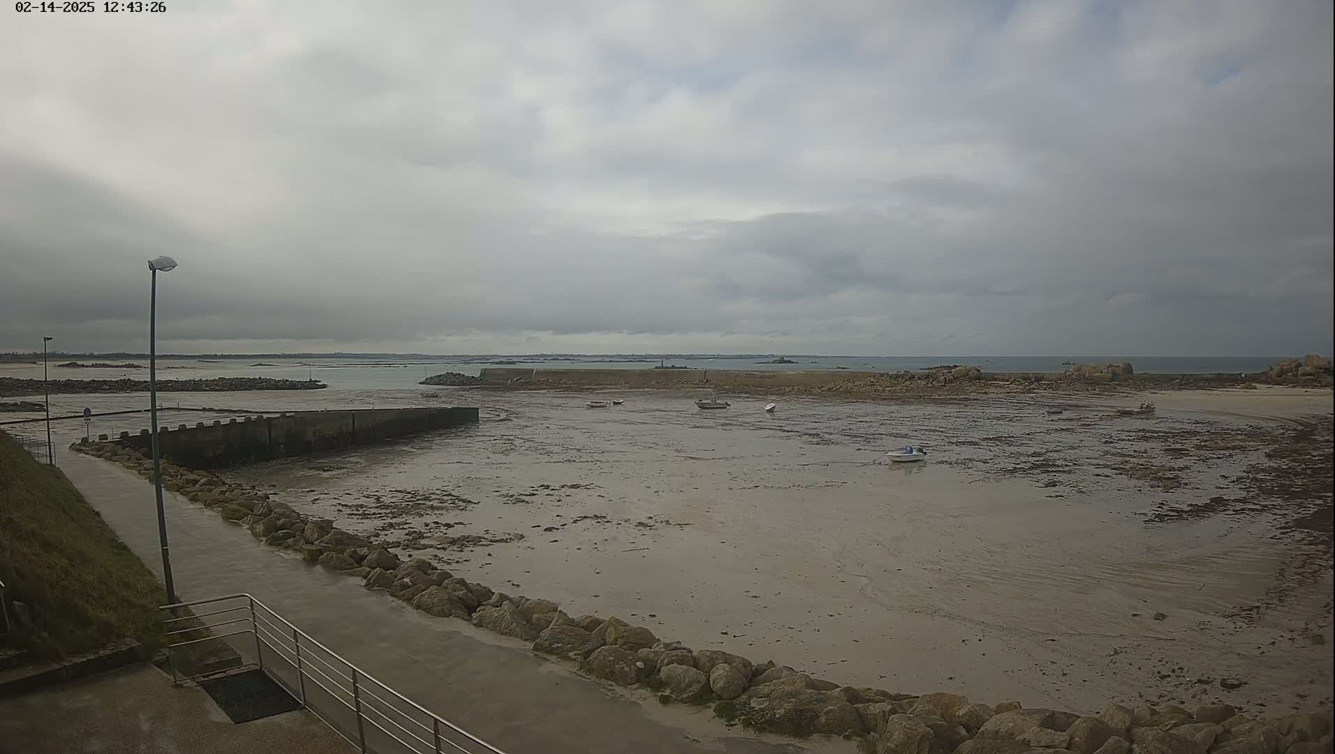 A mostly cloudy day reveals a harbor with several small boats resting on exposed mudflats, surrounded by rocks and a walkway.