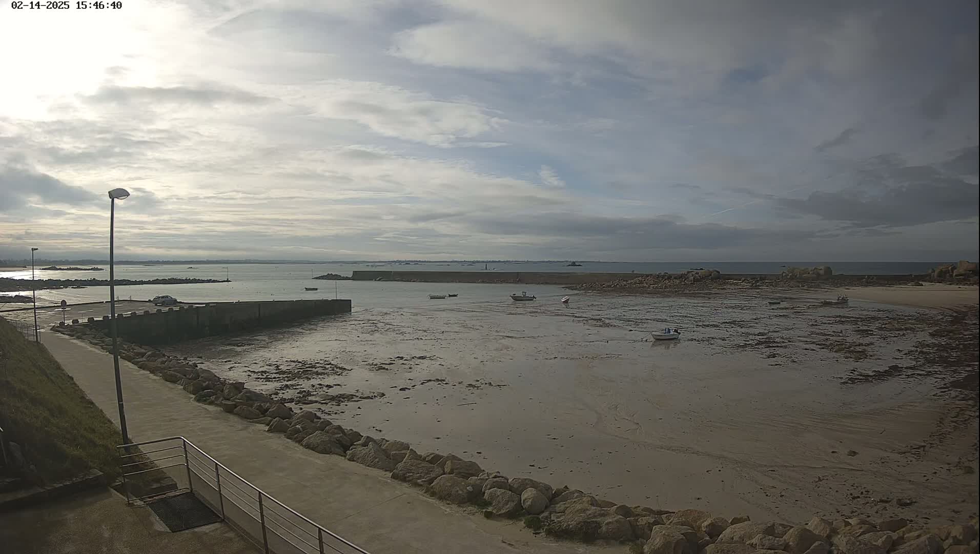 A mostly cloudy day reveals a harbor with several small boats at low tide, a walkway leading to a parking lot, and a rocky shoreline extending into calm water.