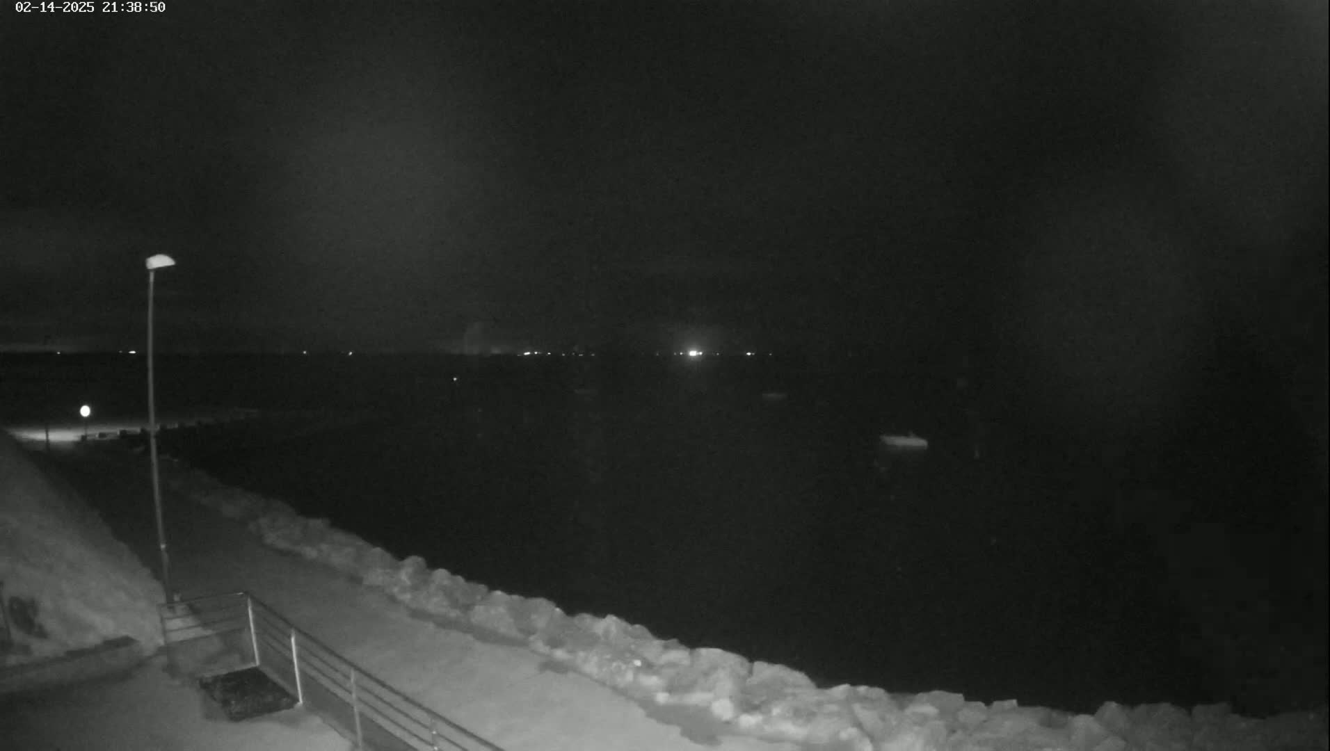 A nighttime view of a dark body of water, possibly a harbor, with a snow-covered walkway and rock barrier in the foreground under a clear, dark sky.