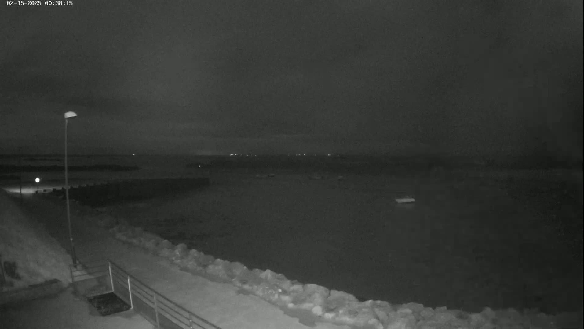 A nighttime view of a calm body of water, possibly a harbor, with a paved walkway and a metal railing leading down to the water's edge, under a dark, cloudy sky.