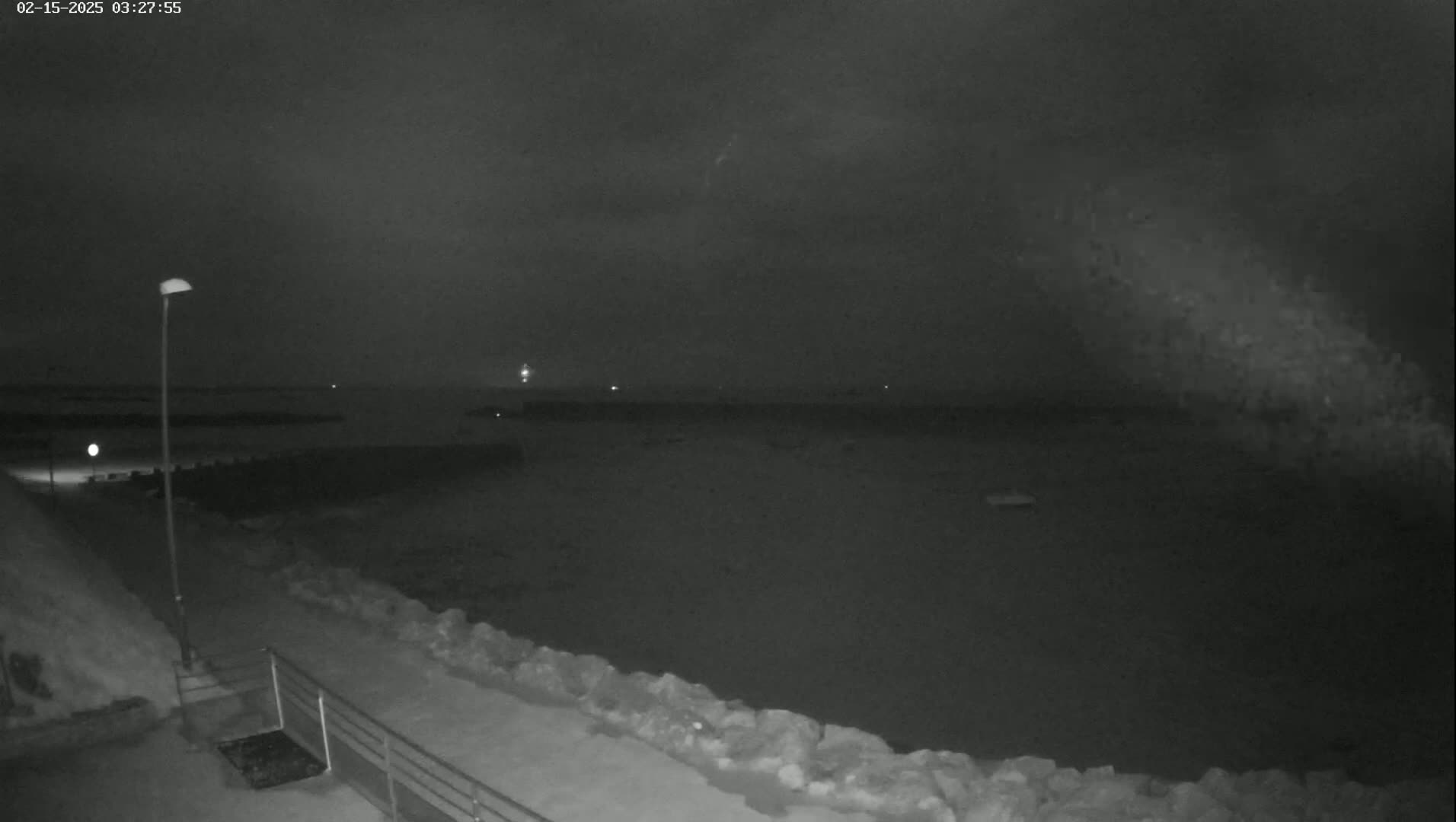 A nighttime, low-light view of a snow-covered shoreline with a calm body of water beyond a low rock wall, under a dark, cloudy sky.