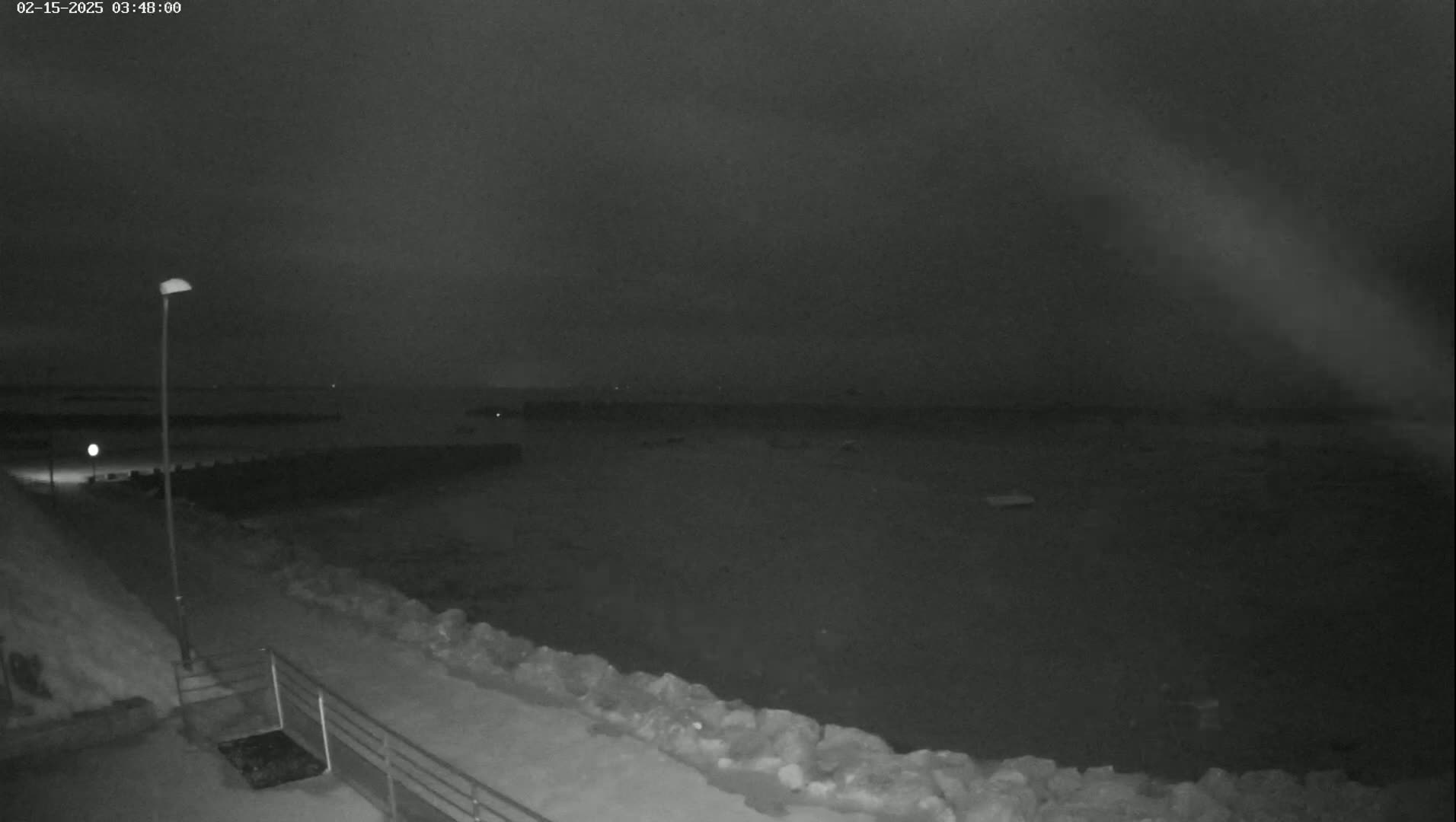 A nighttime, low-light view of a dark, calm body of water with a rocky shoreline and a single lamppost visible along a path.