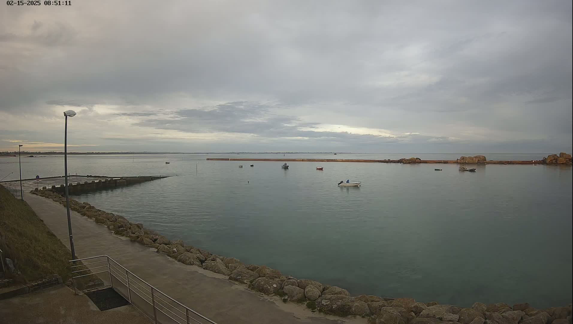 A calm, overcast day shows a harbor with several small boats, a long breakwater, and a paved walkway leading down to the water's edge.