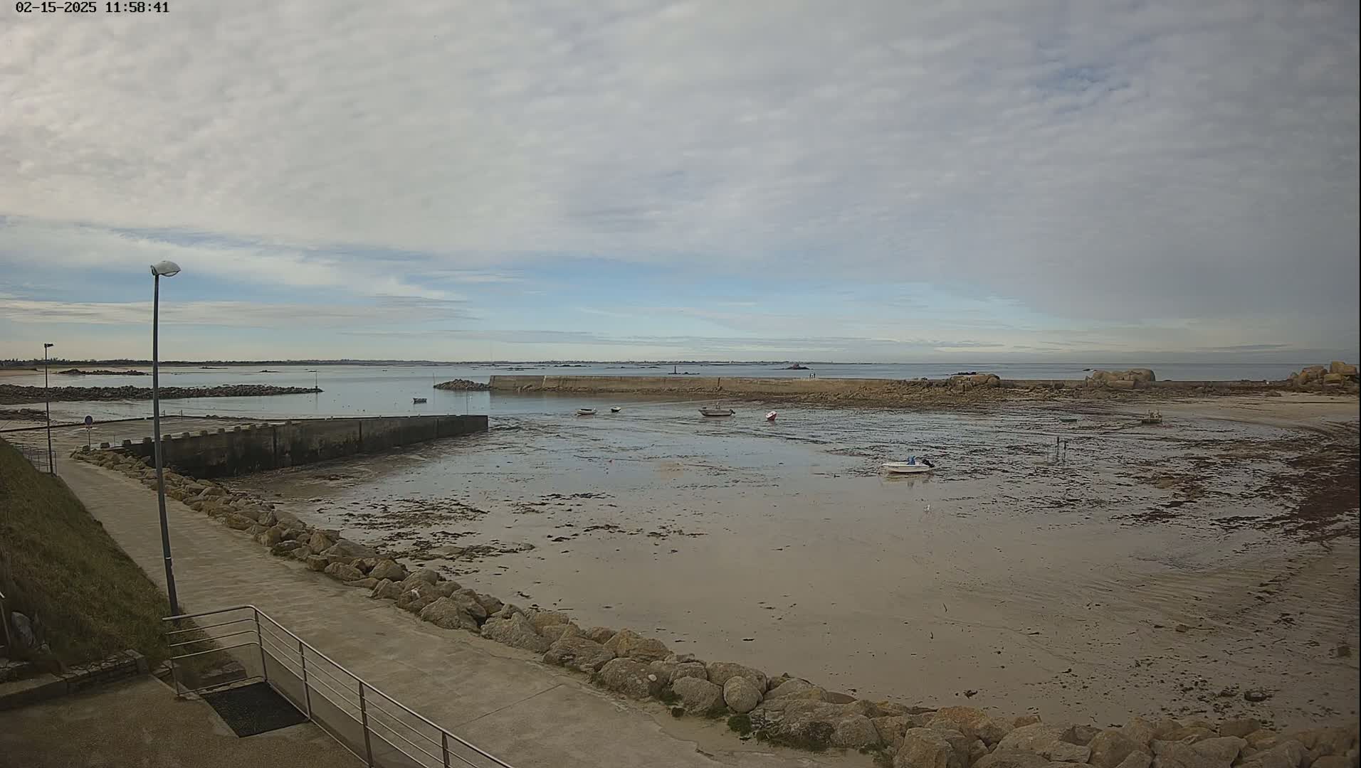 A mostly cloudy day reveals a small harbor with several small boats at low tide, a concrete walkway, and a rocky shoreline extending to a calm sea.