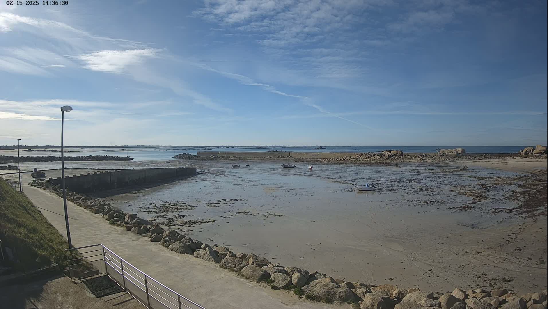 A mostly sunny day reveals a harbor with a few small boats at low tide, a paved walkway leading to the water's edge, and rocky areas surrounding the sandy expanse.