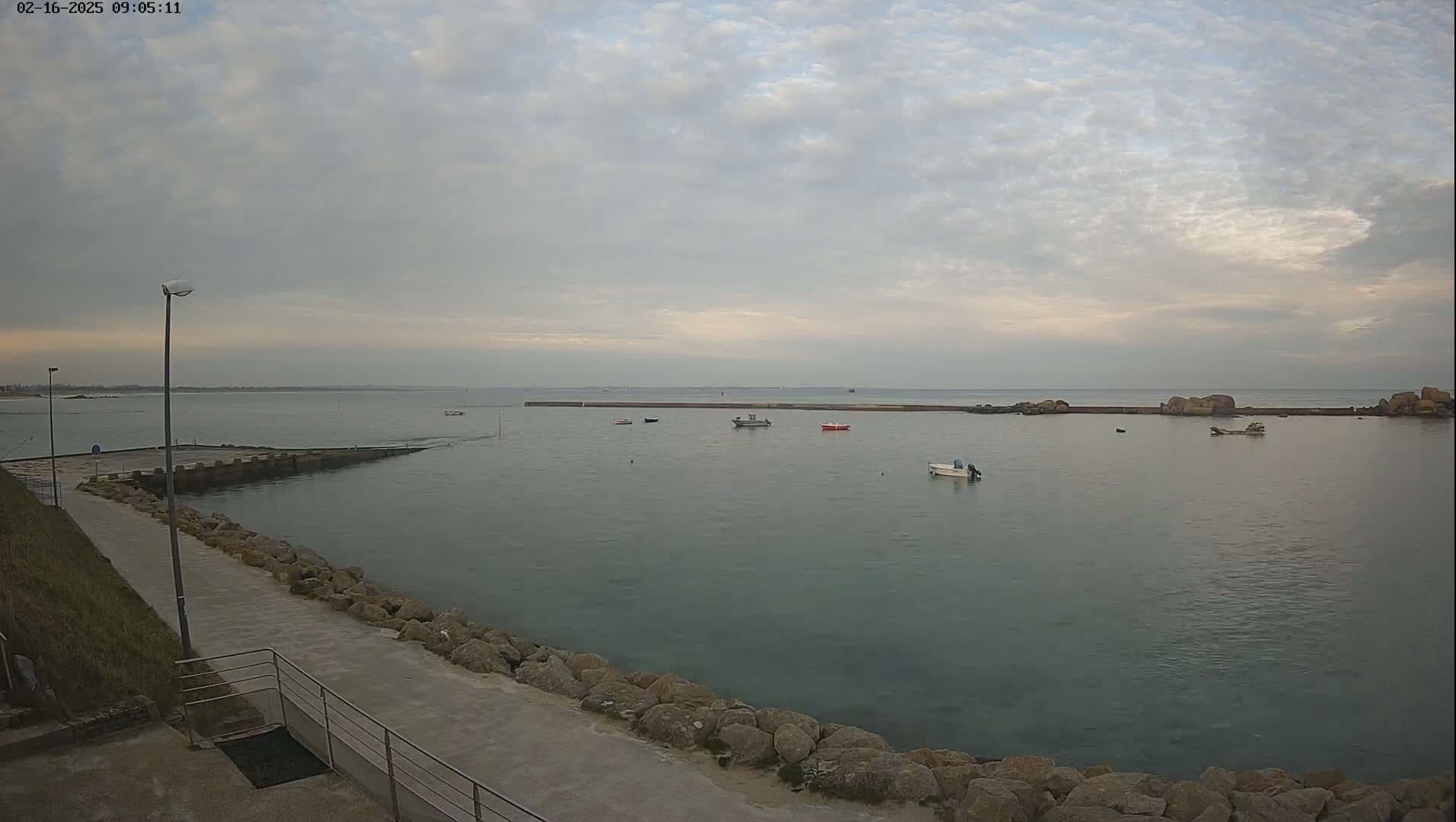 A calm body of water with several small boats is seen from a paved walkway lined with rocks under a cloudy sky.