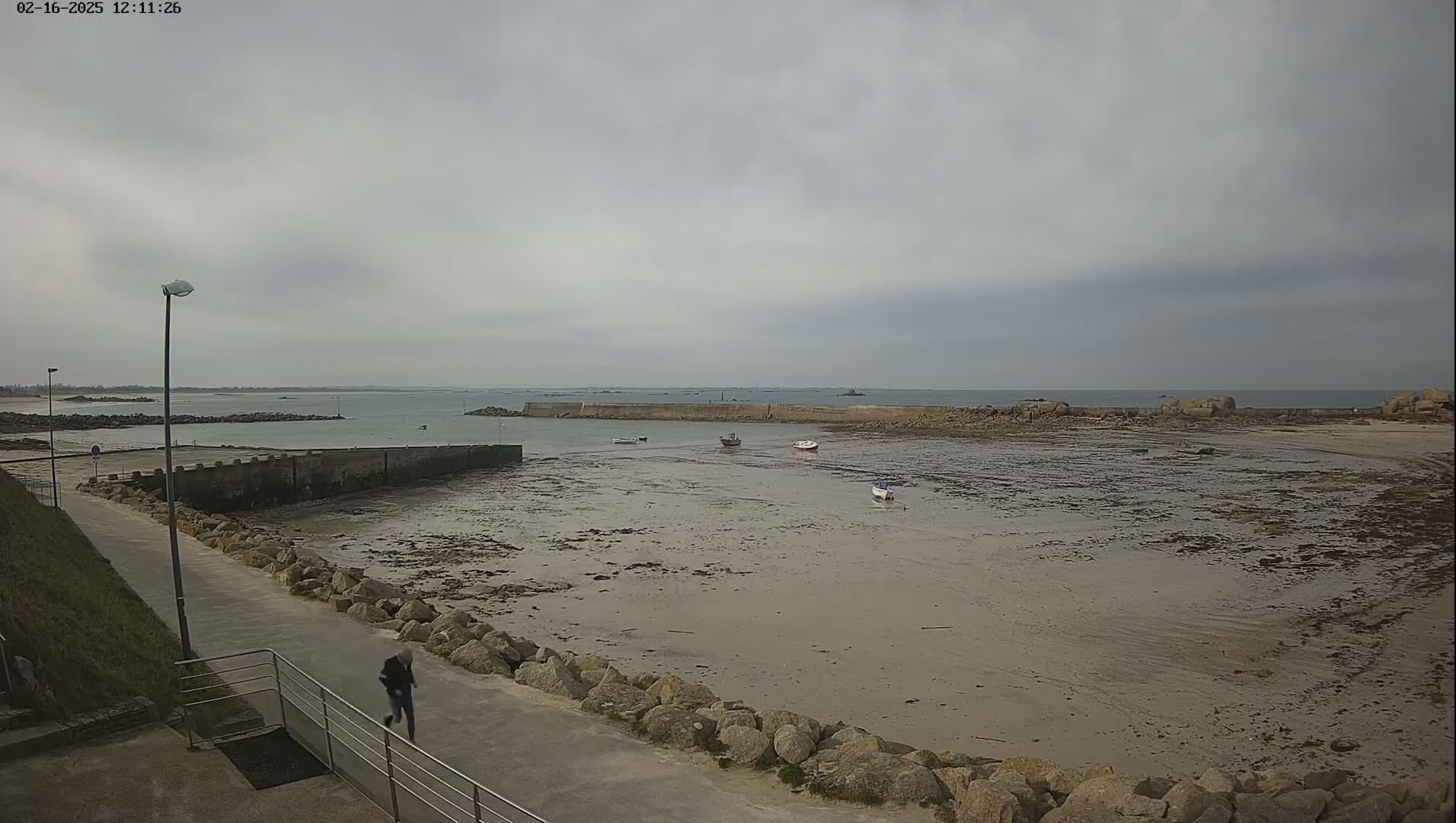 A mostly overcast day reveals a rocky coastline with a small harbor containing several small boats at low tide, and a person walking on a paved path nearby.