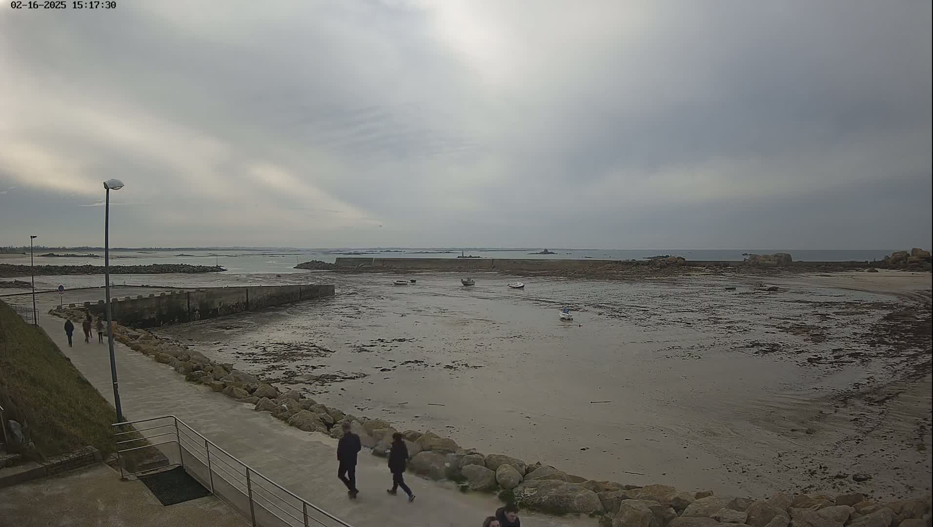 A mostly cloudy day reveals a rocky, low-tide harbor with several small boats and a few people walking along a paved path.