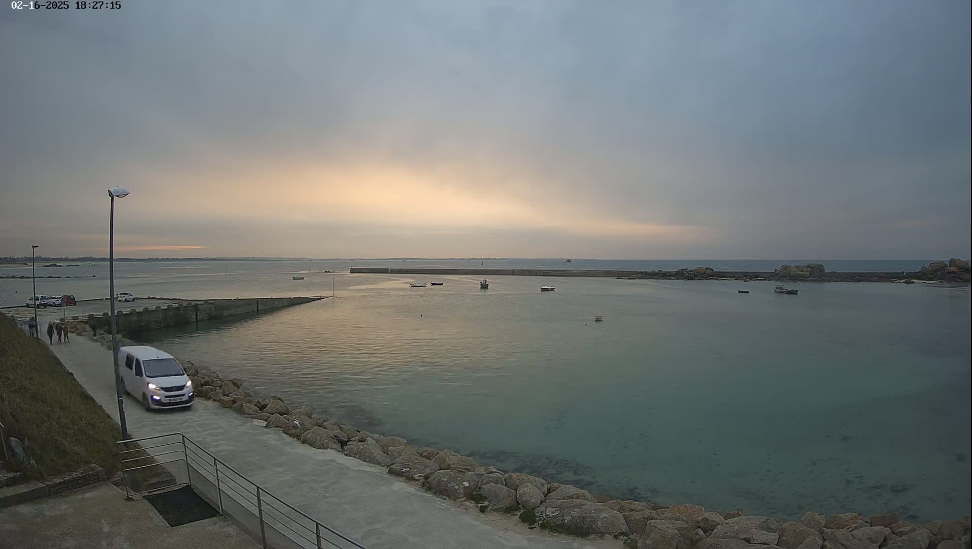 A calm, pale-sunset-lit harbor with several small boats, a white van parked on a paved road beside a rocky shoreline.