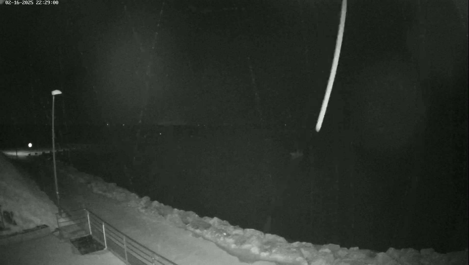 A nighttime, snowy scene shows a bright streak in the sky above a snow-covered waterfront path with a lamppost.
