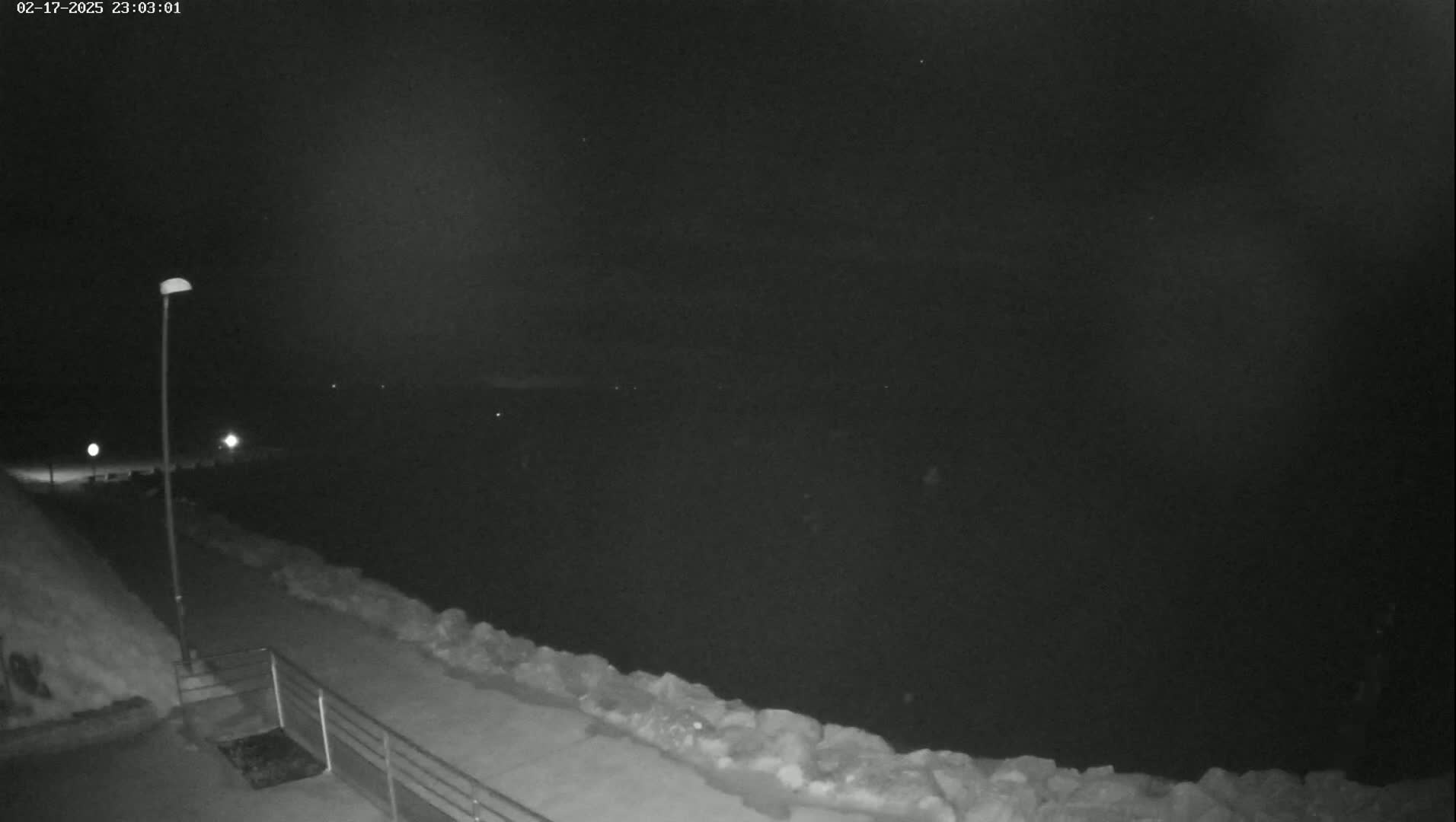 A nighttime, low-light view shows a snow-covered walkway next to a dark body of water under a mostly clear sky.
