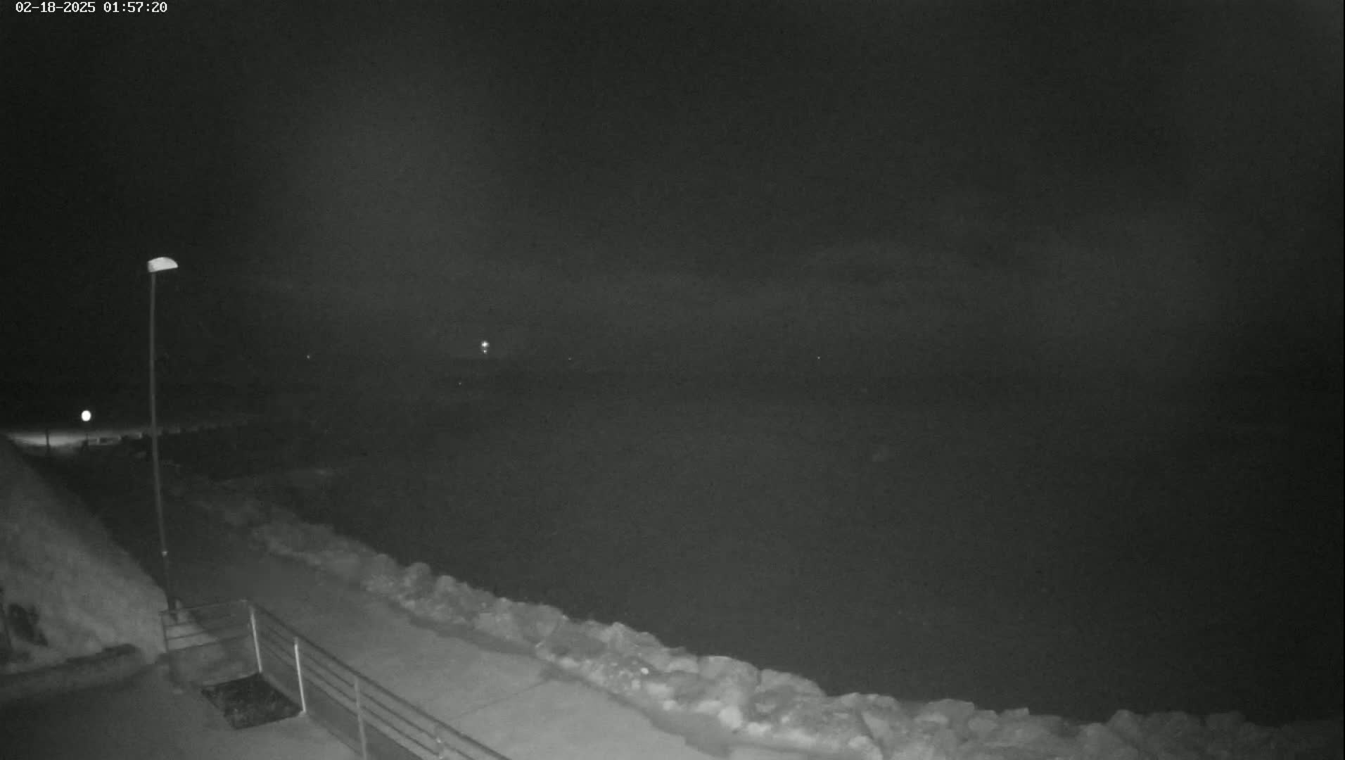 A nighttime, low-light view of a snow-covered walkway next to a dark body of water, illuminated by a single lamppost.