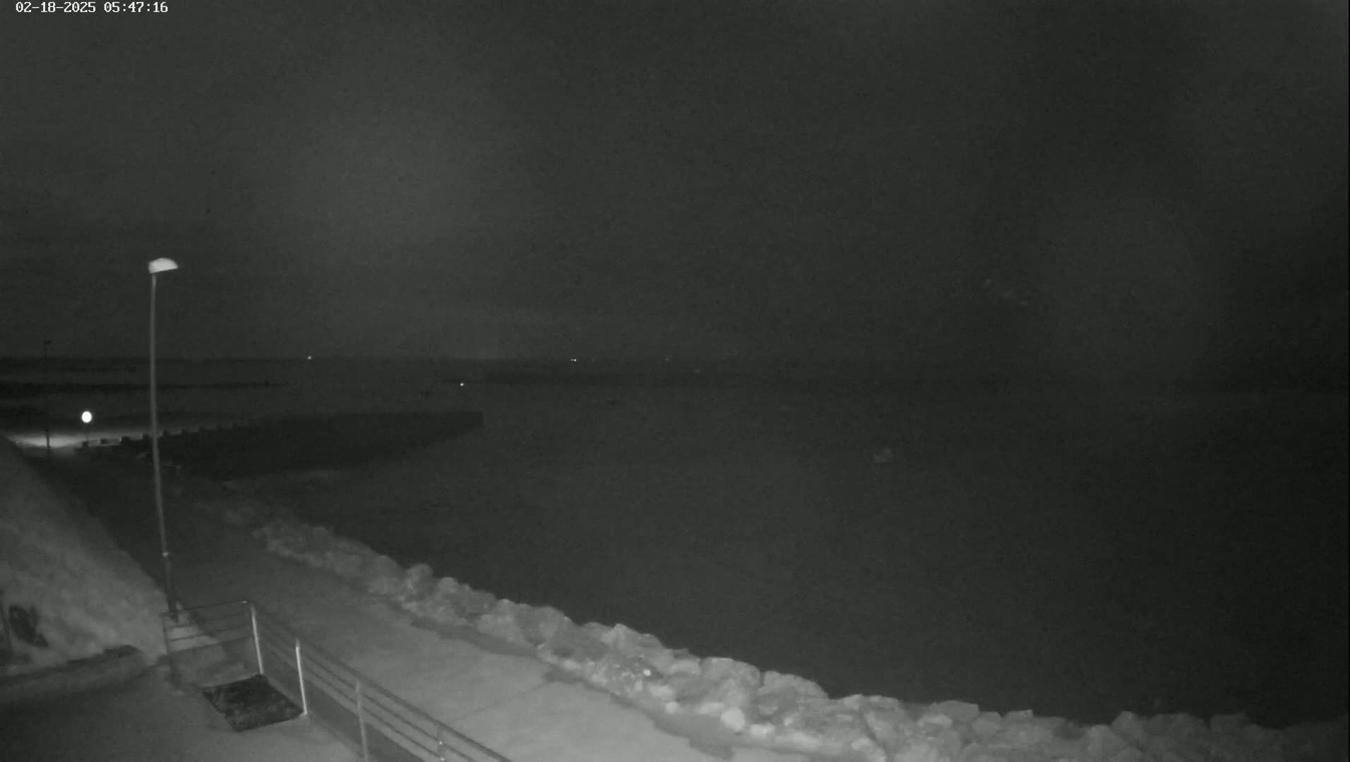 A nighttime, snow-covered waterfront scene shows a dark body of water, a lamppost, and a walkway bordered by a low rock wall.