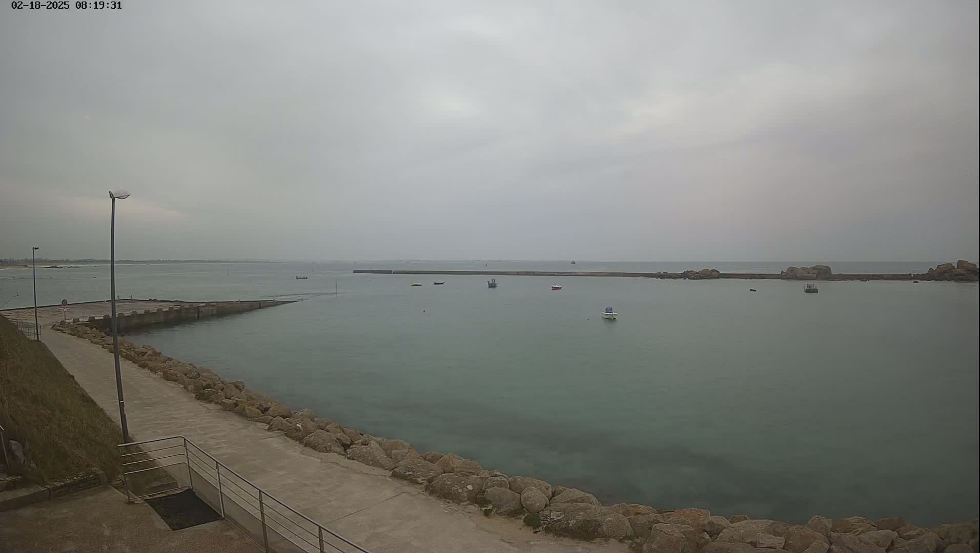 A calm, overcast day reveals a harbor with several small boats, a concrete walkway, and rocky shorelines.