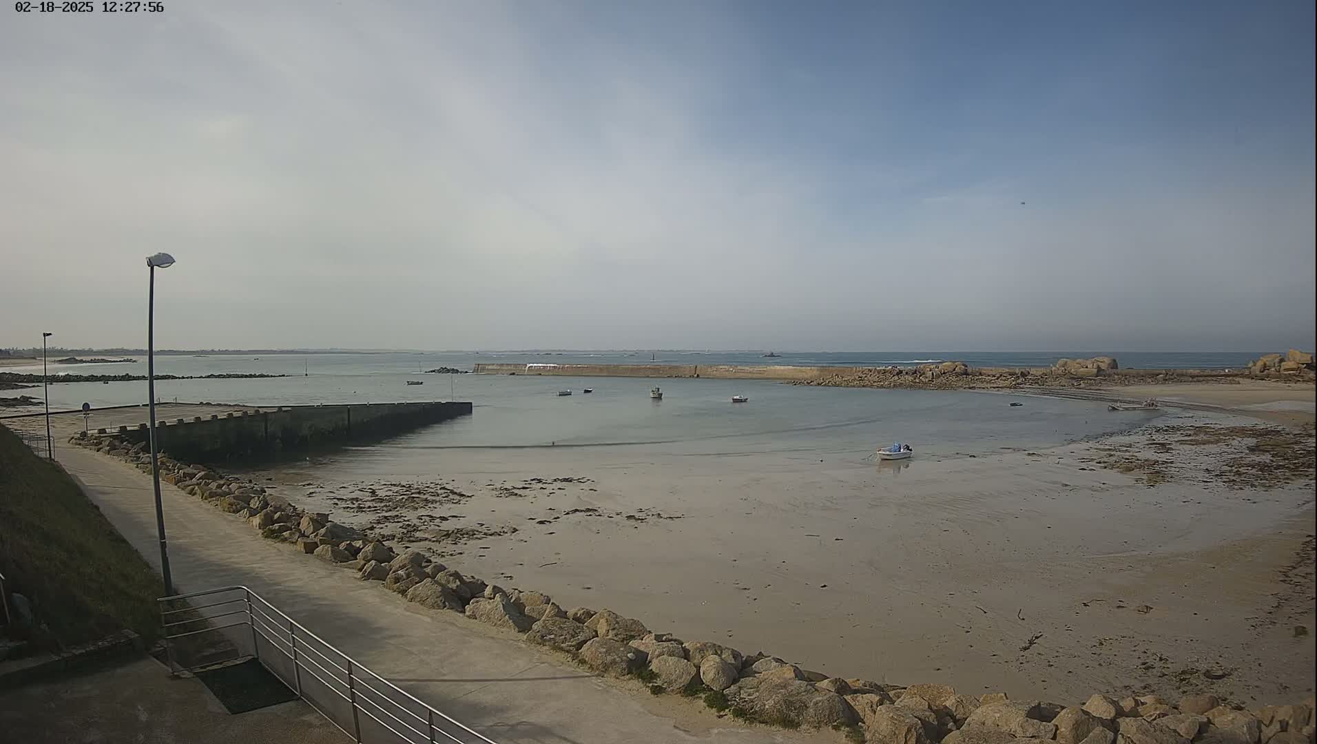 A mostly sunny day shows a rocky, sandy cove with several small boats, a low stone wall, and a paved walkway leading to it.