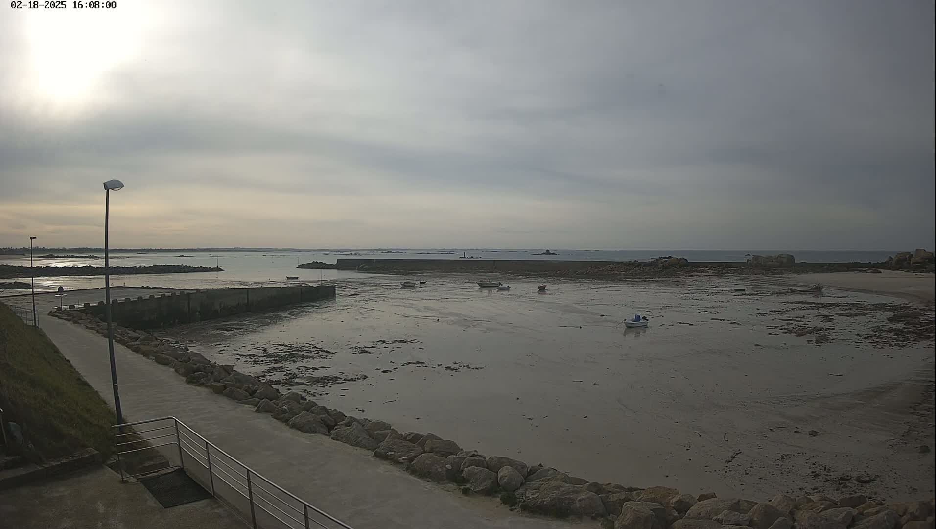 A mostly cloudy day reveals a low-tide harbor with several small boats resting on exposed mudflats, a concrete seawall, and a paved walkway leading to the water's edge.