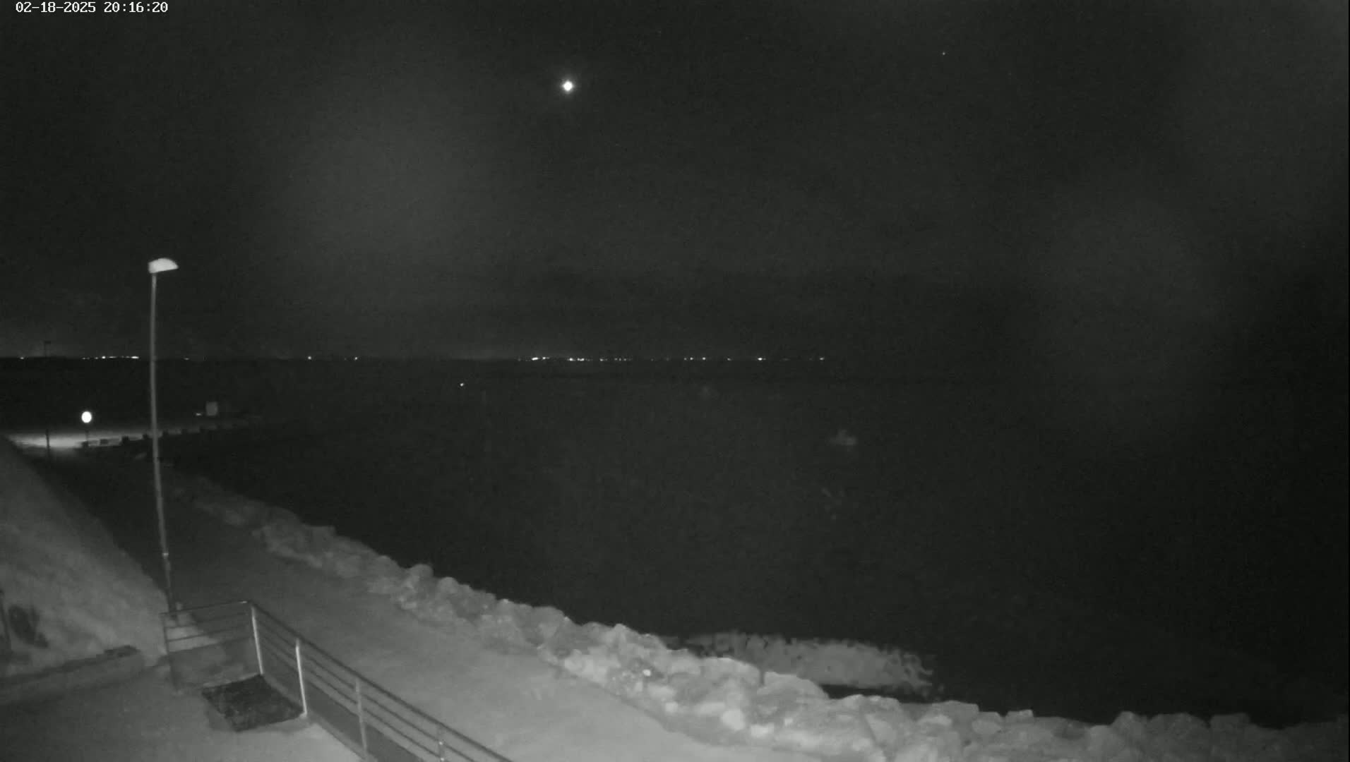 A nighttime, snowy scene shows a body of water with distant lights, viewed from a snow-covered walkway with a lamppost and railing.