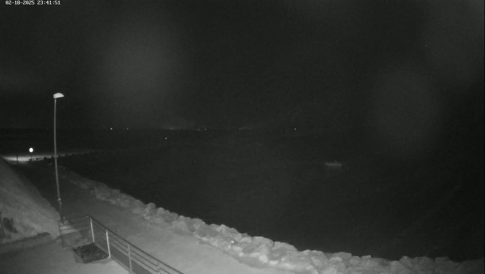 A nighttime, low-light view shows a snow-covered path along a rocky shoreline, with a single lamppost illuminating the foreground.