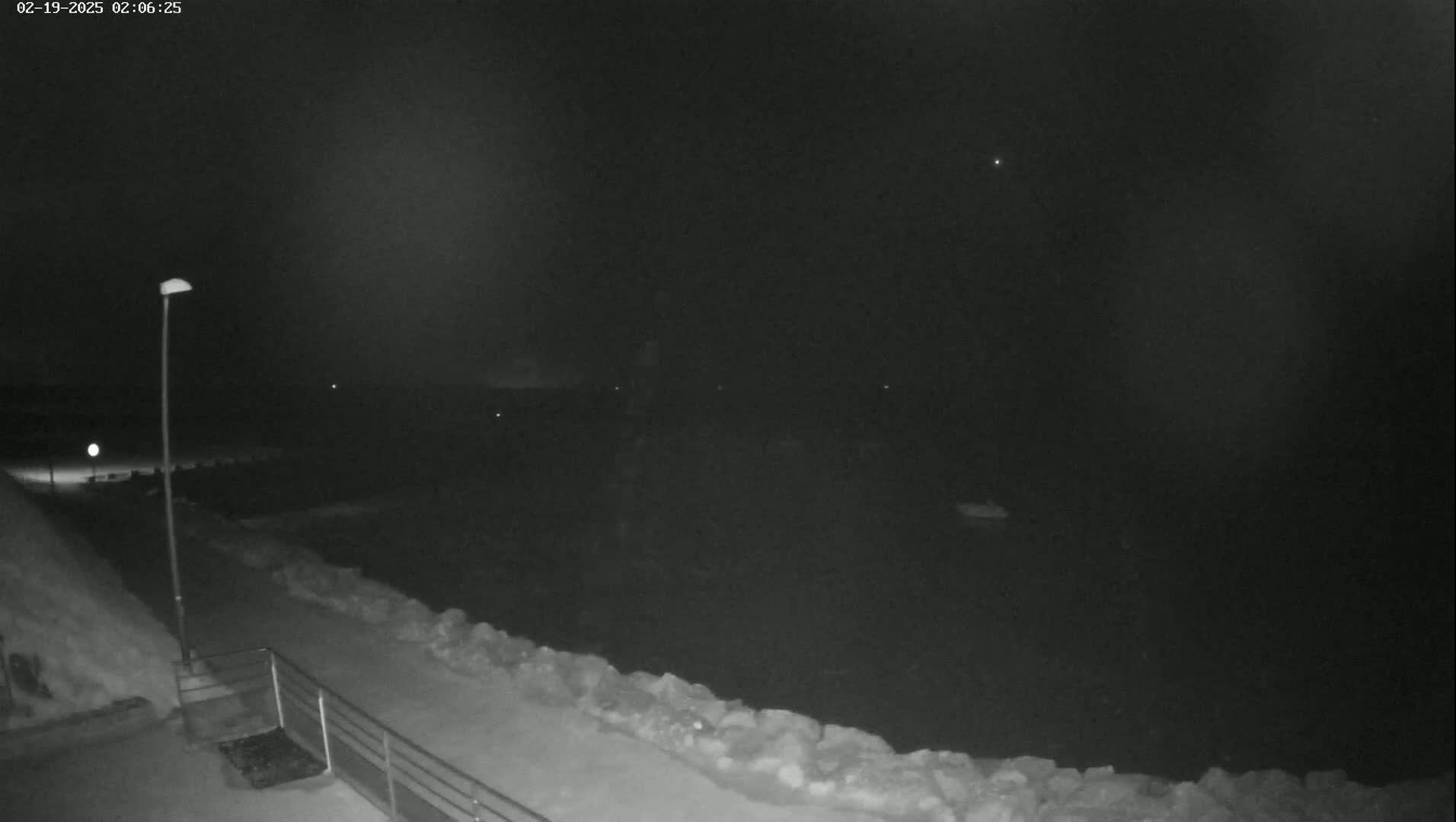 A nighttime, snowy scene shows a dark body of water, a snow-covered walkway with a railing, and a single lit lamppost.