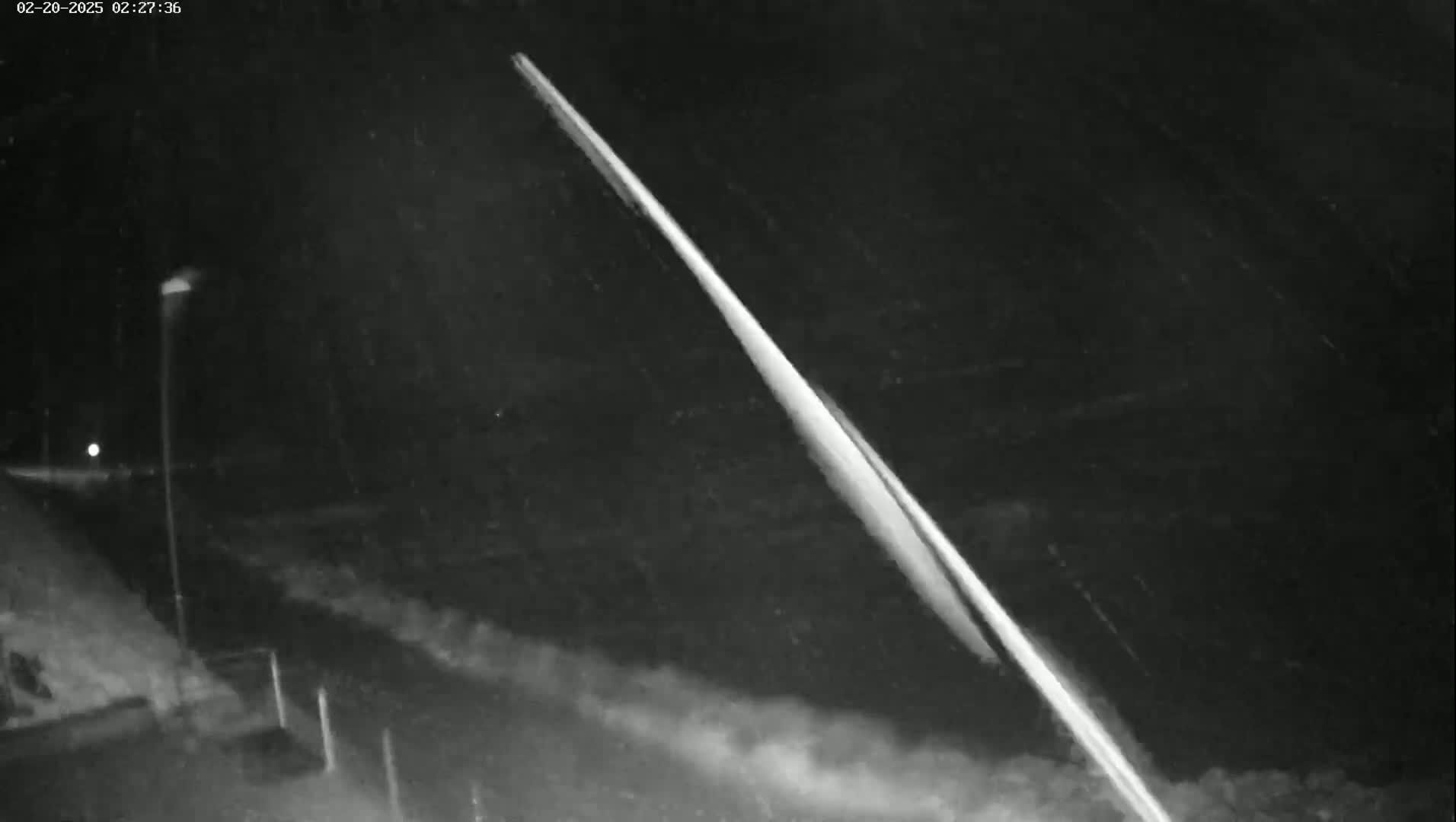 A bright streak of light diagonally crosses a dark, snowy nighttime scene, illuminated by a single streetlamp.