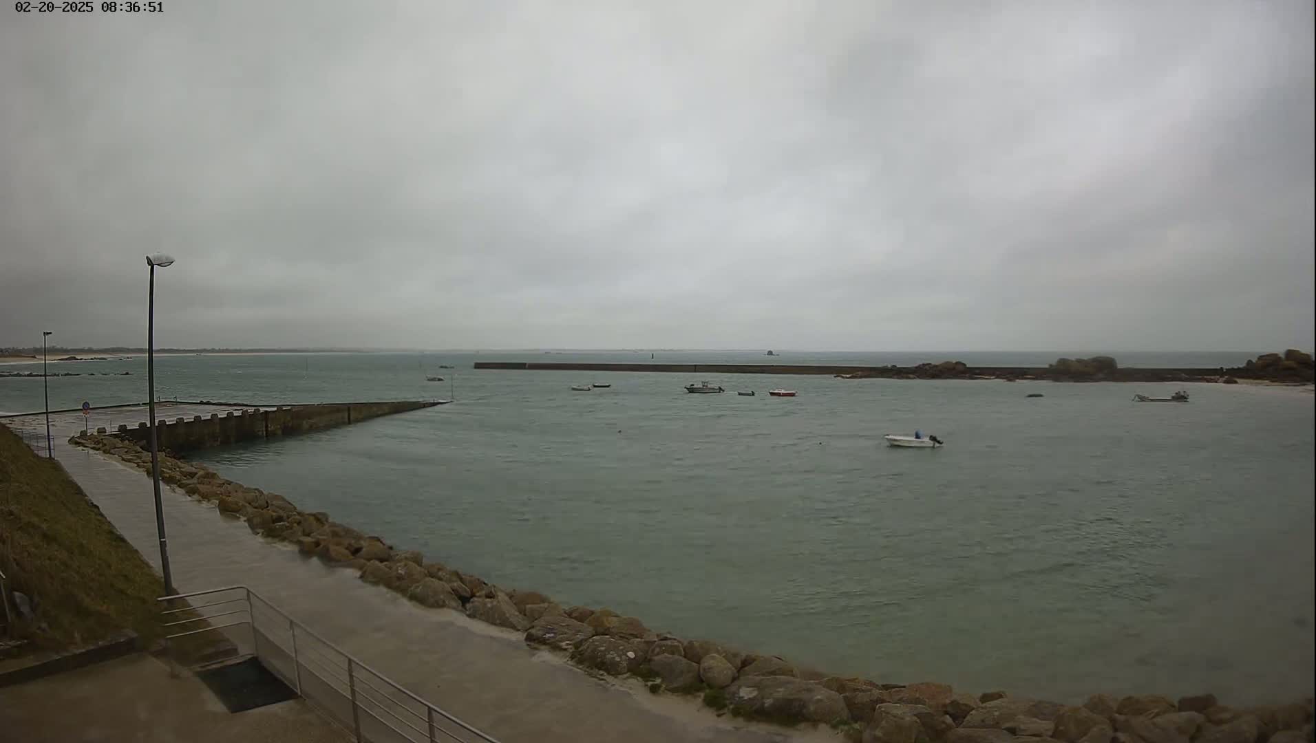 A calm, overcast day shows a harbor with several small boats, a long jetty, and rocky shoreline.