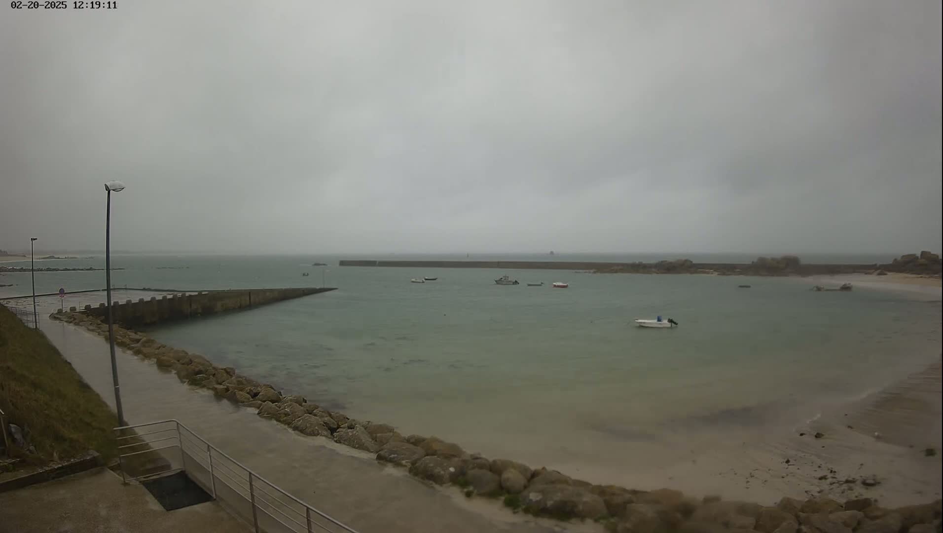 A calm, overcast day shows a small harbor with several boats, a concrete walkway, and rocky shores.